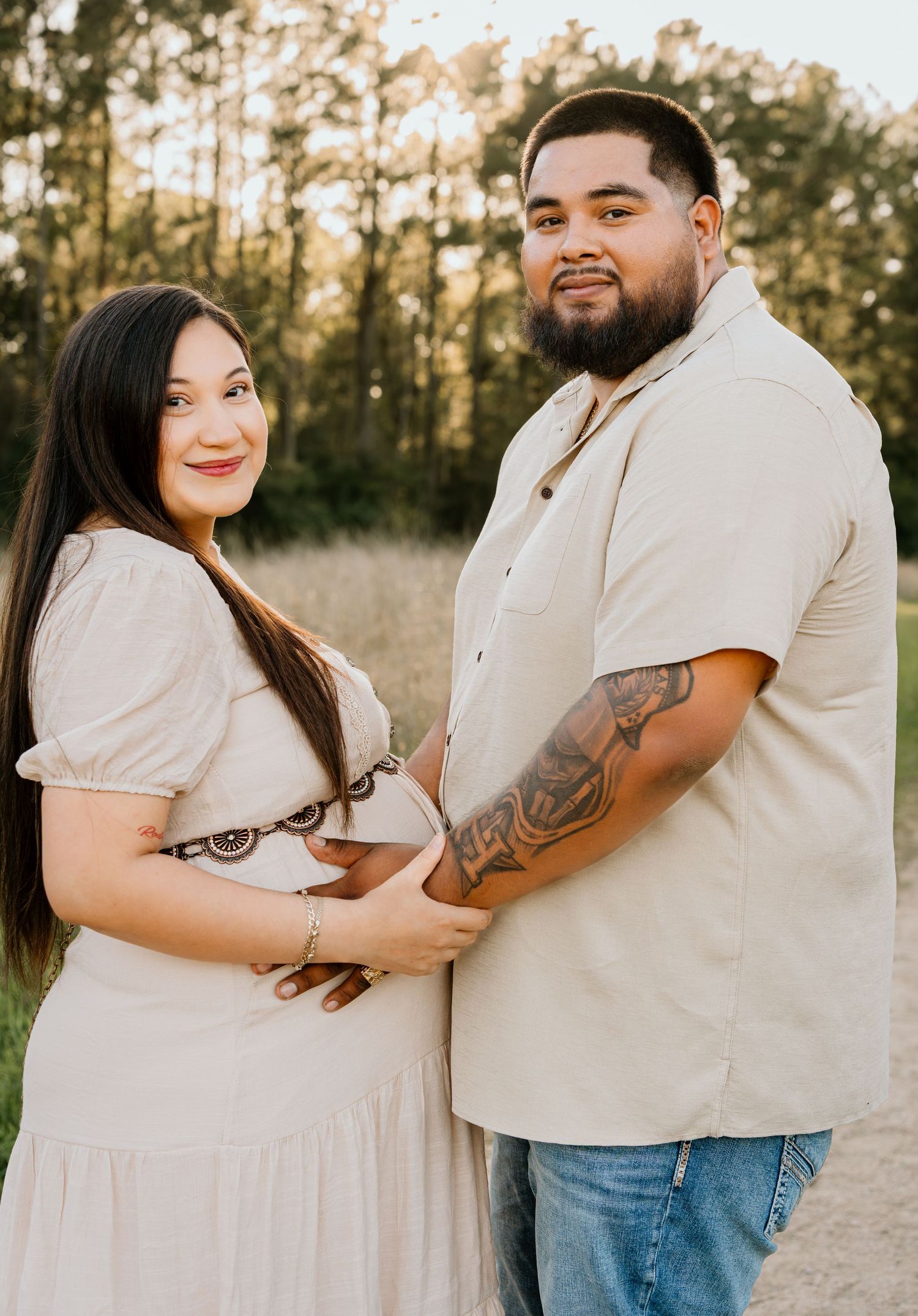 A couple poses outdoors during a maternity photo shoot, the woman in a white dress cradling her baby bump.