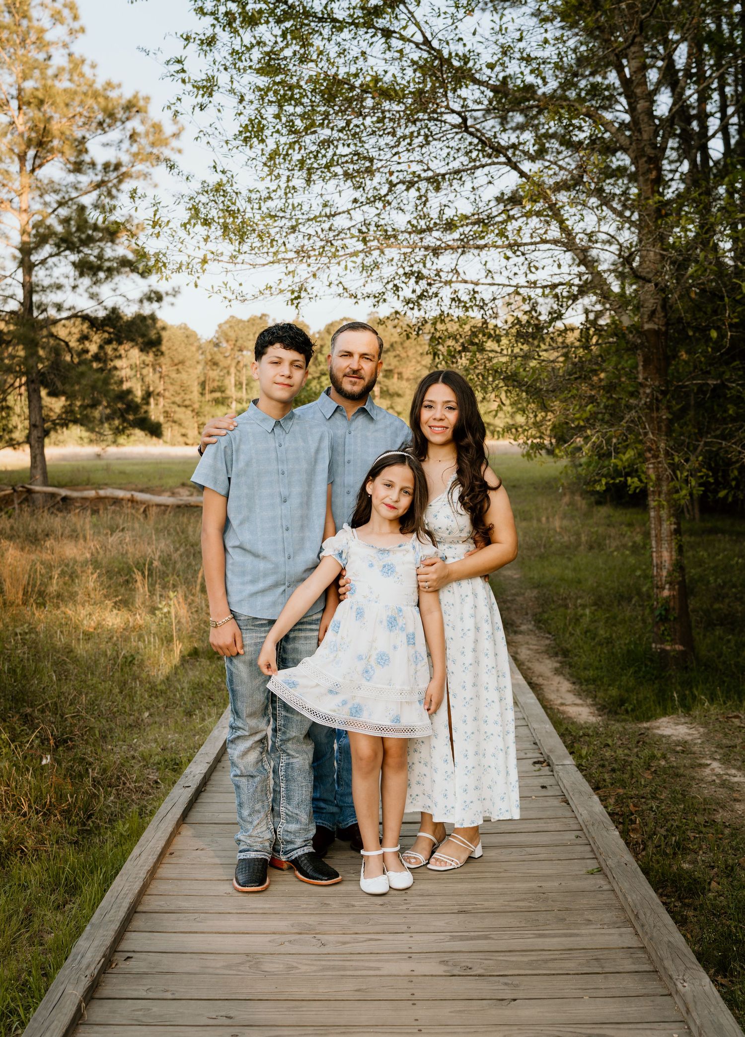 Family of four posing on a wooden boardwalk path surrounded by trees and golden meadow during an outdoor portrait session.