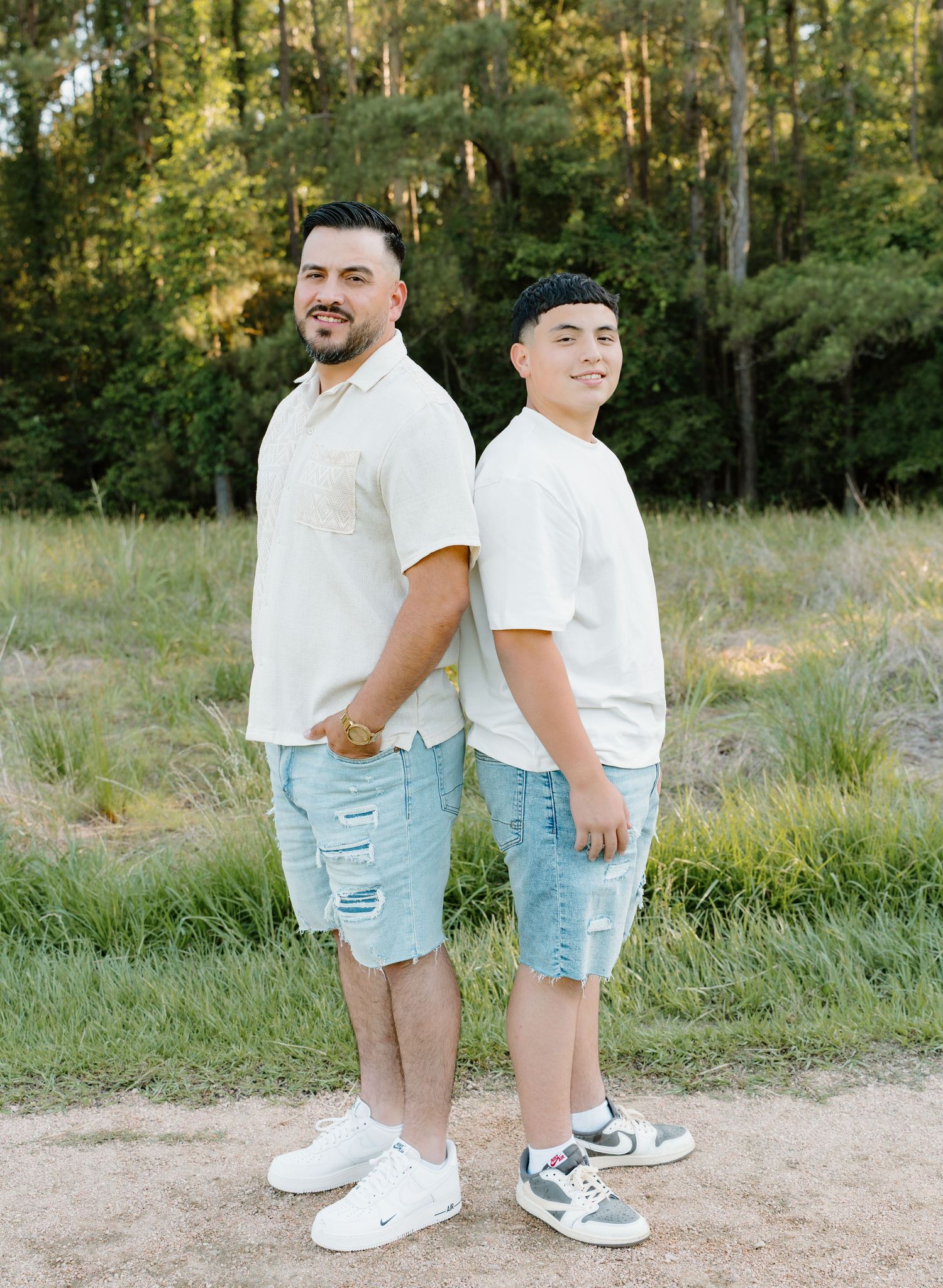 Two young men in light shirts and denim shorts pose back-to-back outdoors near a grassy field.