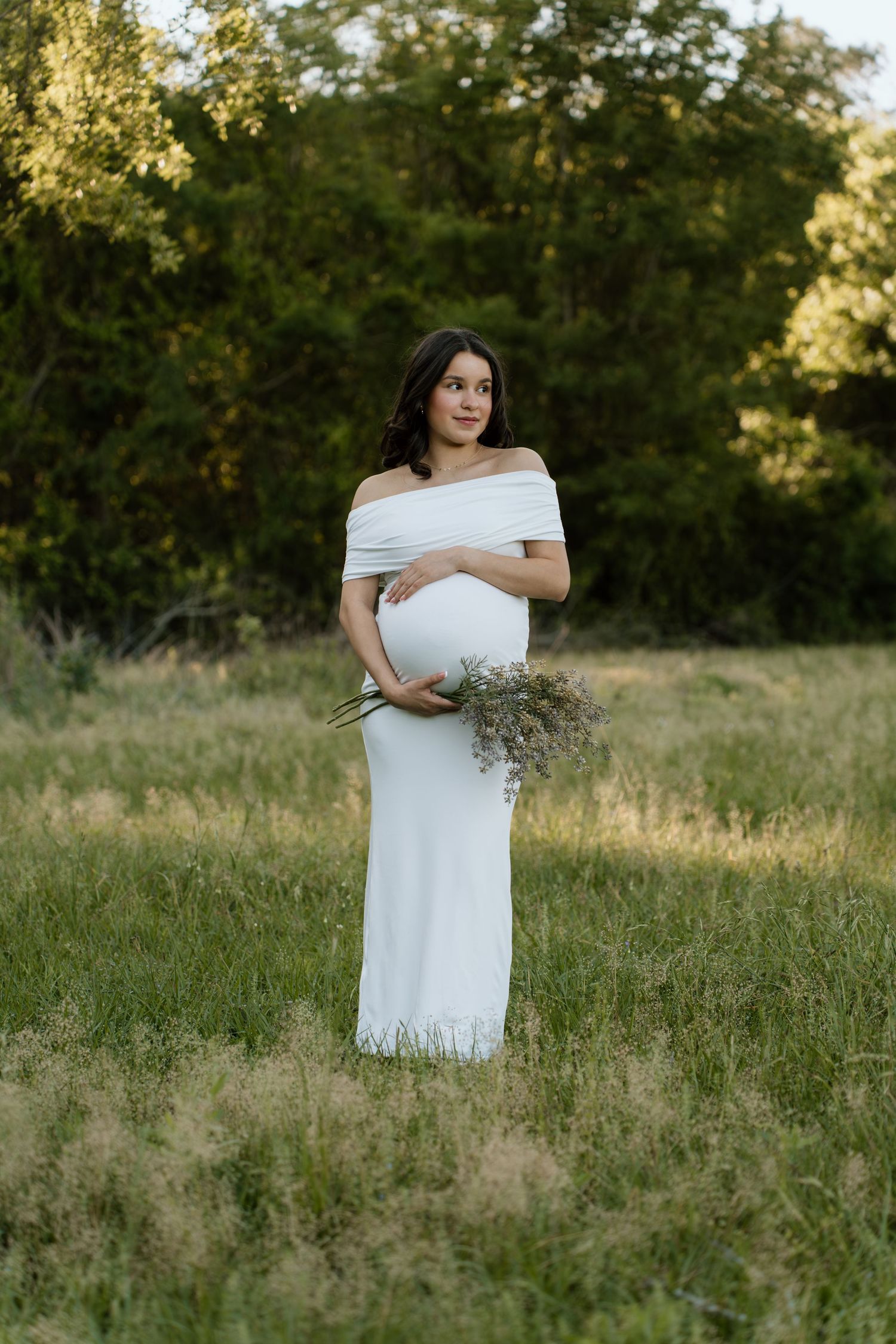 A pregnant woman in a white off-shoulder gown holds wildflowers while standing in a sunlit meadow.