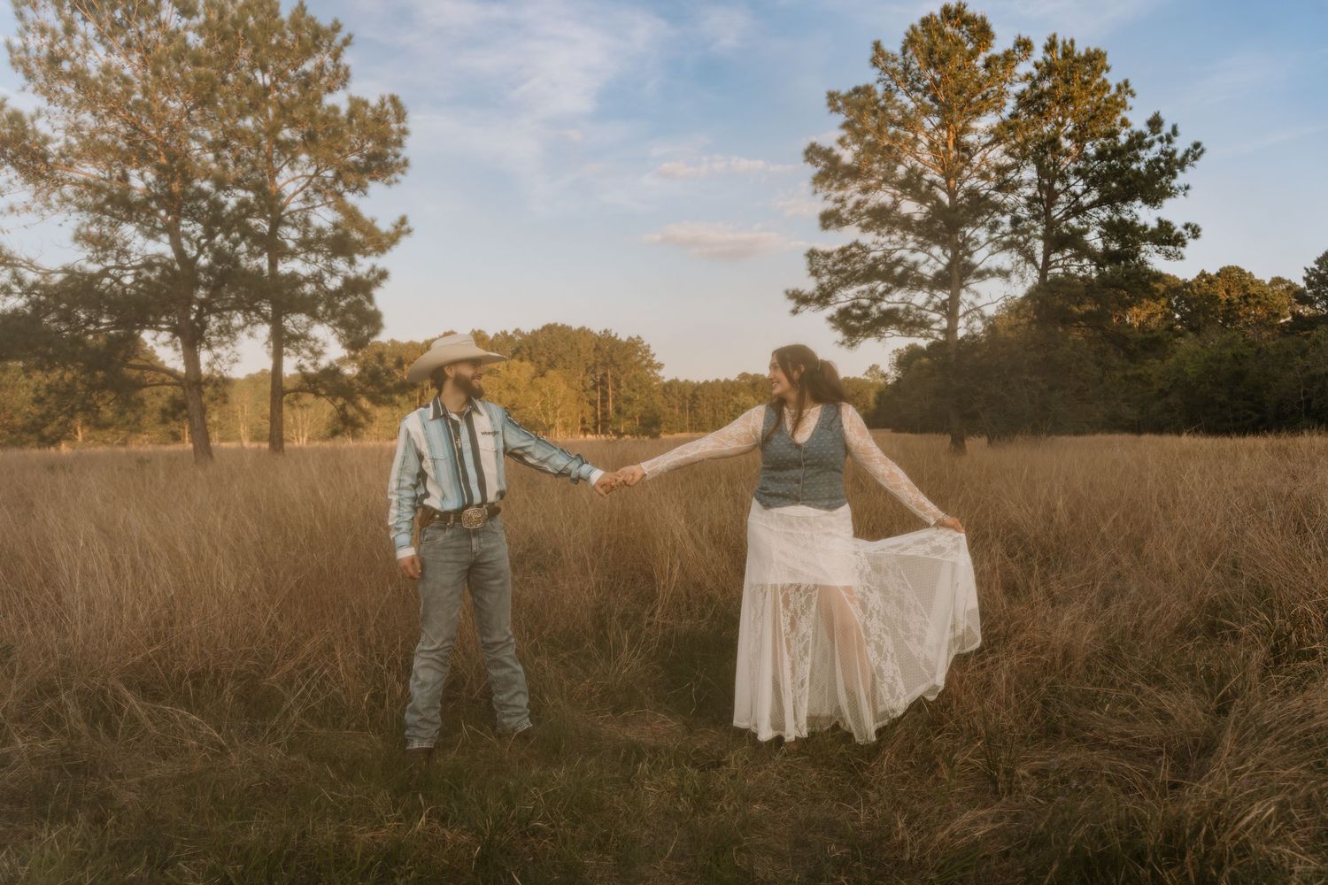 Couple holding hands in golden field at sunset, man in cowboy hat and woman in white lace skirt among tall grass.