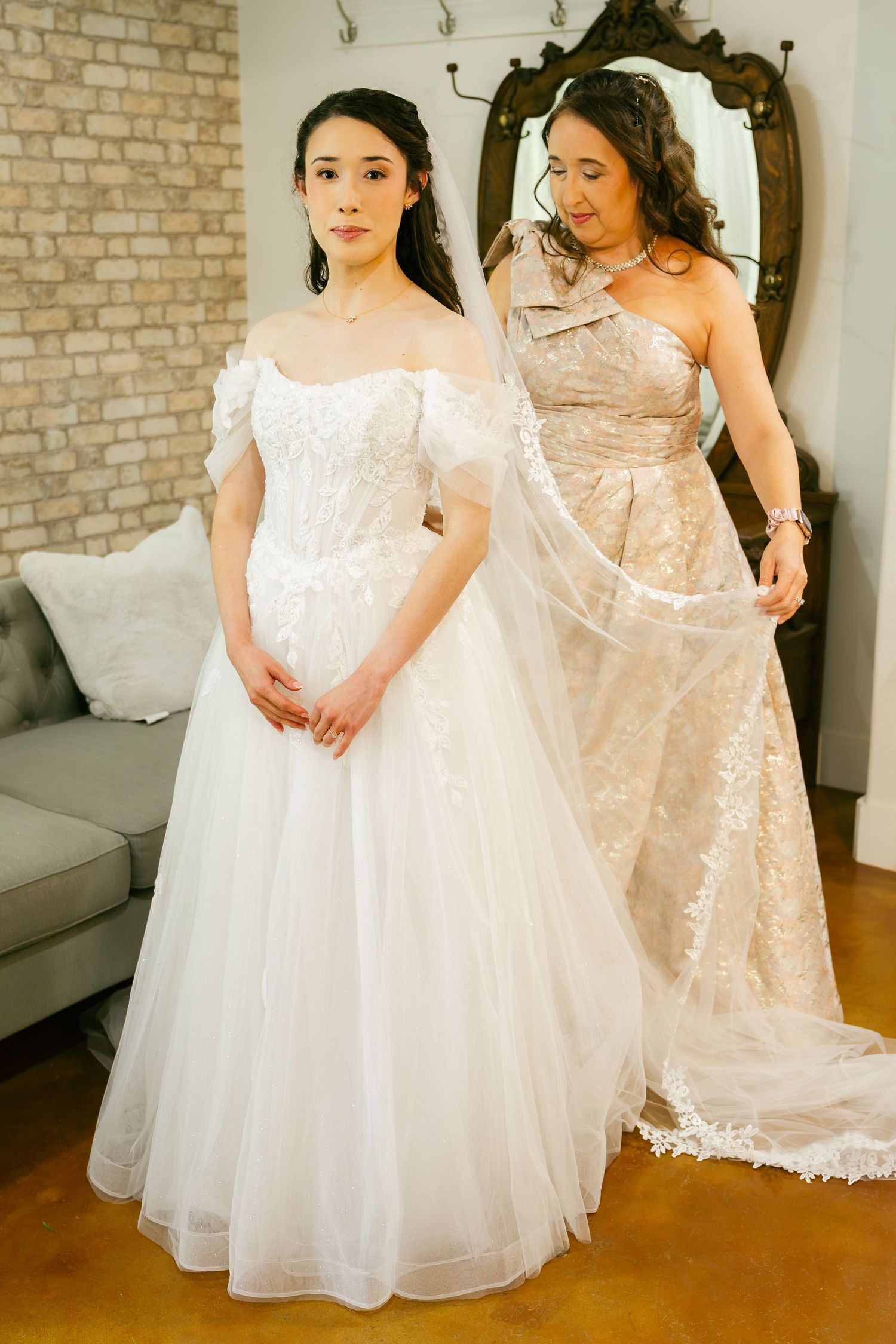 Bride in off-shoulder lace wedding gown being assisted with veil by woman in champagne dress in bridal boutique.