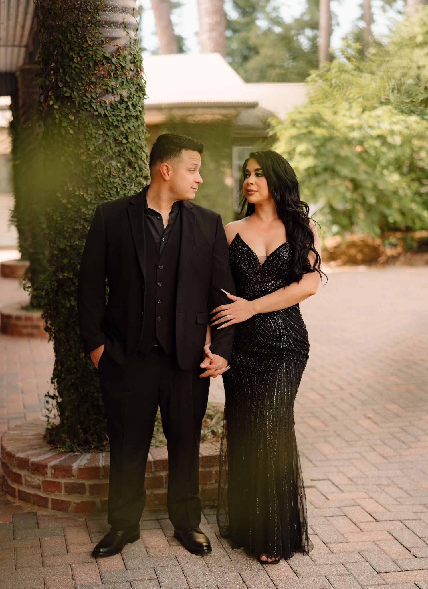 Couple in matching black formal attire posing outdoors on brick pathway surrounded by lush greenery.