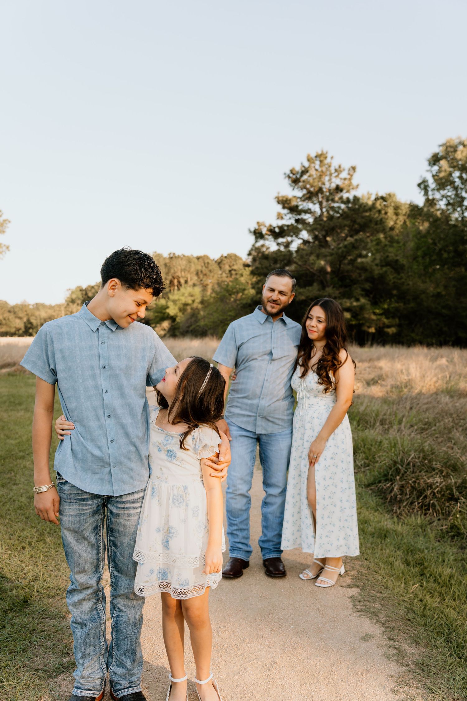 Family of four in coordinating blue and white outfits walking a sunny dirt path surrounded by green trees and golden grass.