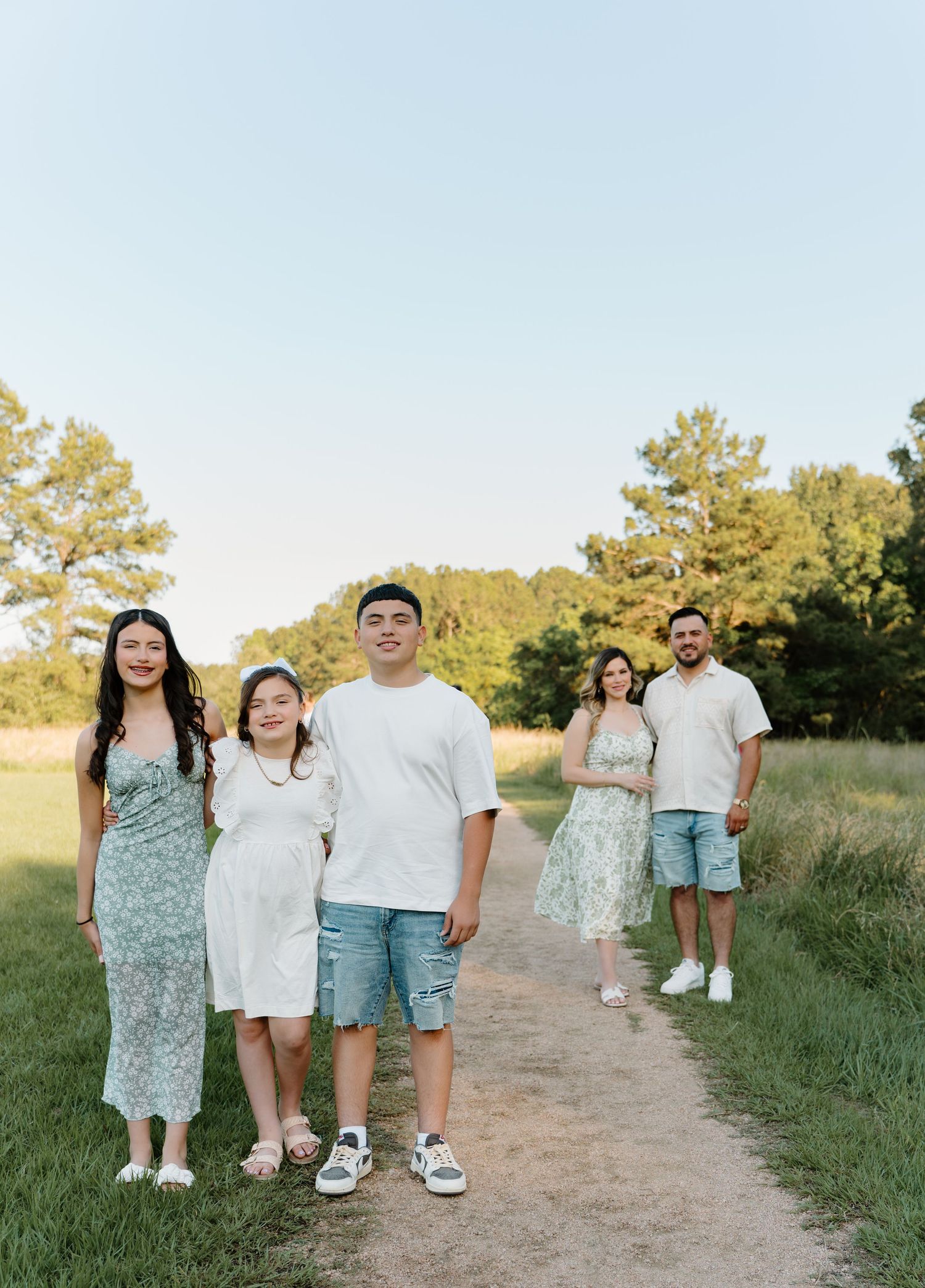 Family of five posing on a scenic dirt path surrounded by green trees and fields during golden hour.