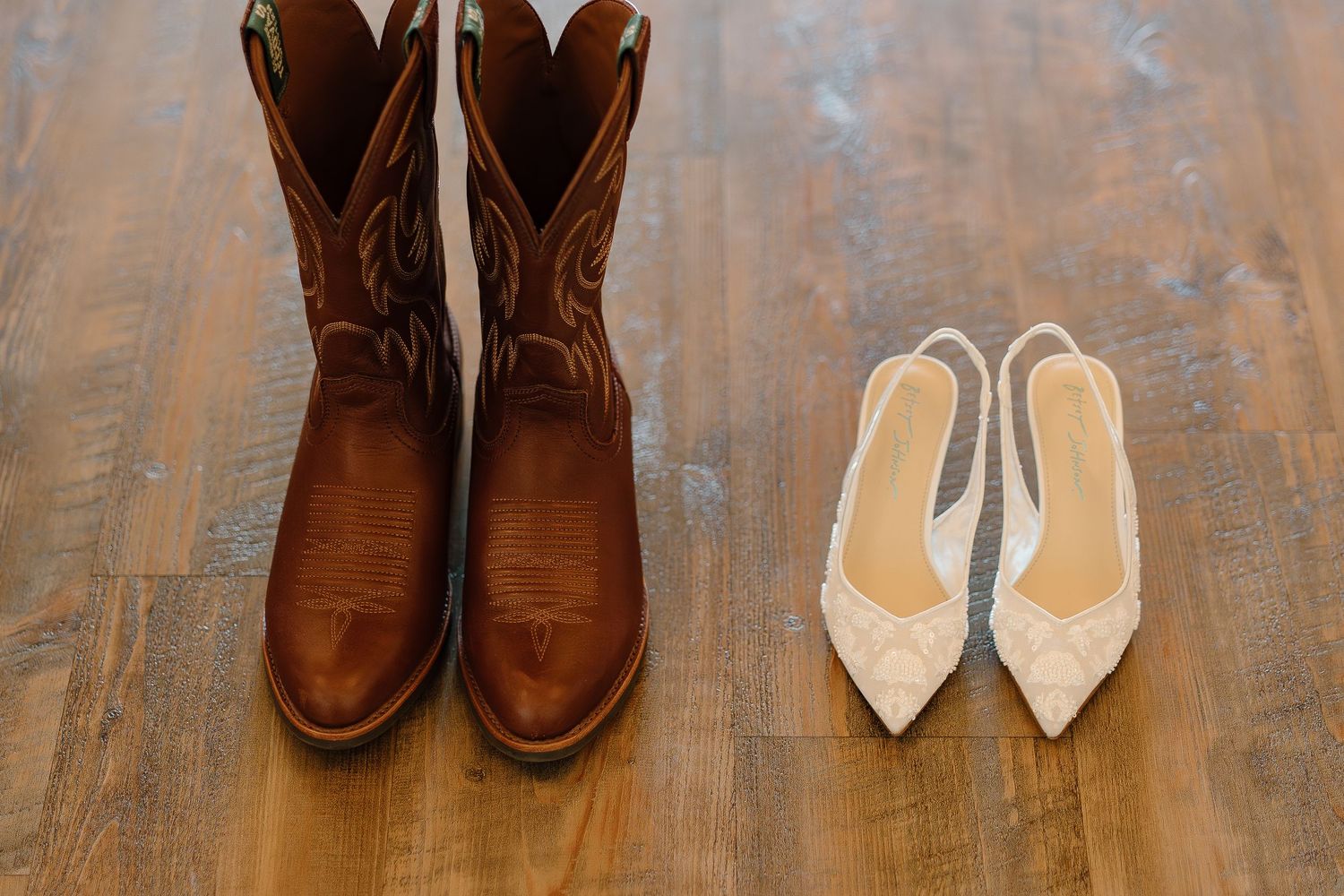 Brown cowboy boots beside white lace bridal heels on rustic wood floor, perfect contrast for country wedding footwear.