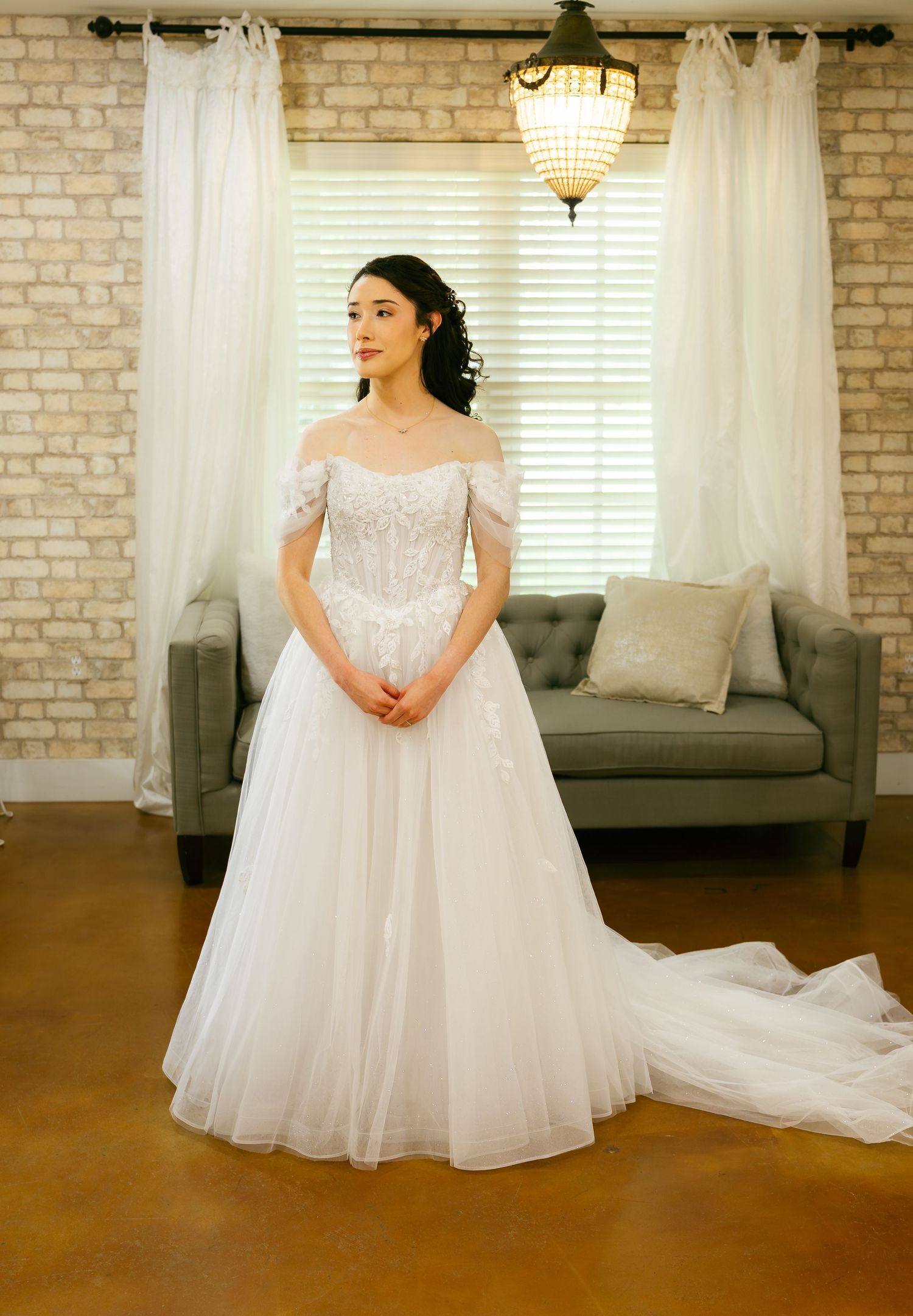 Elegant bride in off-shoulder lace ball gown standing in a bright rustic venue.