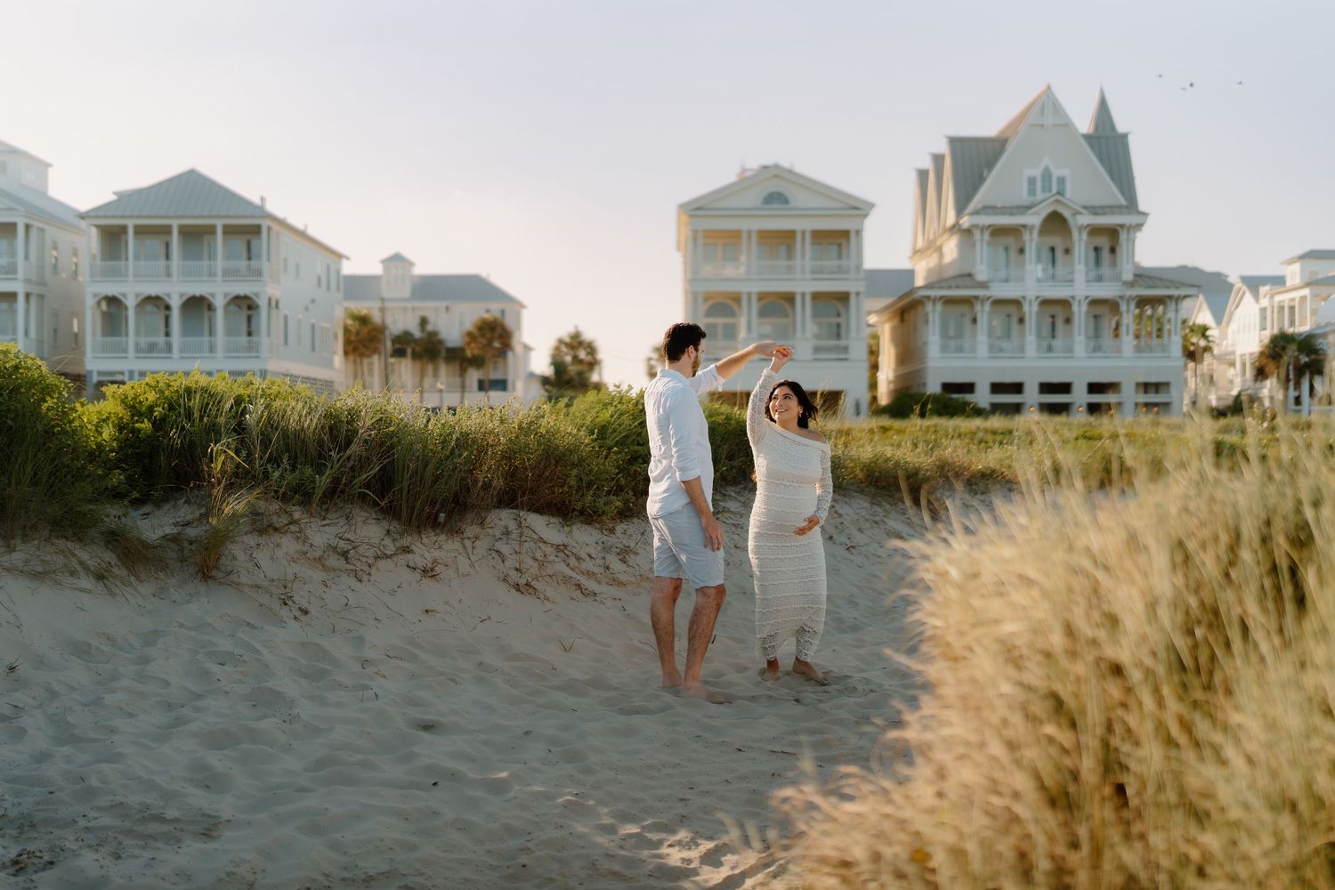 A couple dances barefoot on sandy beach dunes with white coastal vacation homes in the background at golden hour.