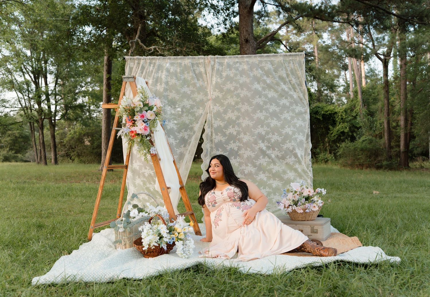 Pregnant woman in pink dress sits on blanket beside floral arch backdrop outdoors for maternity photo.
