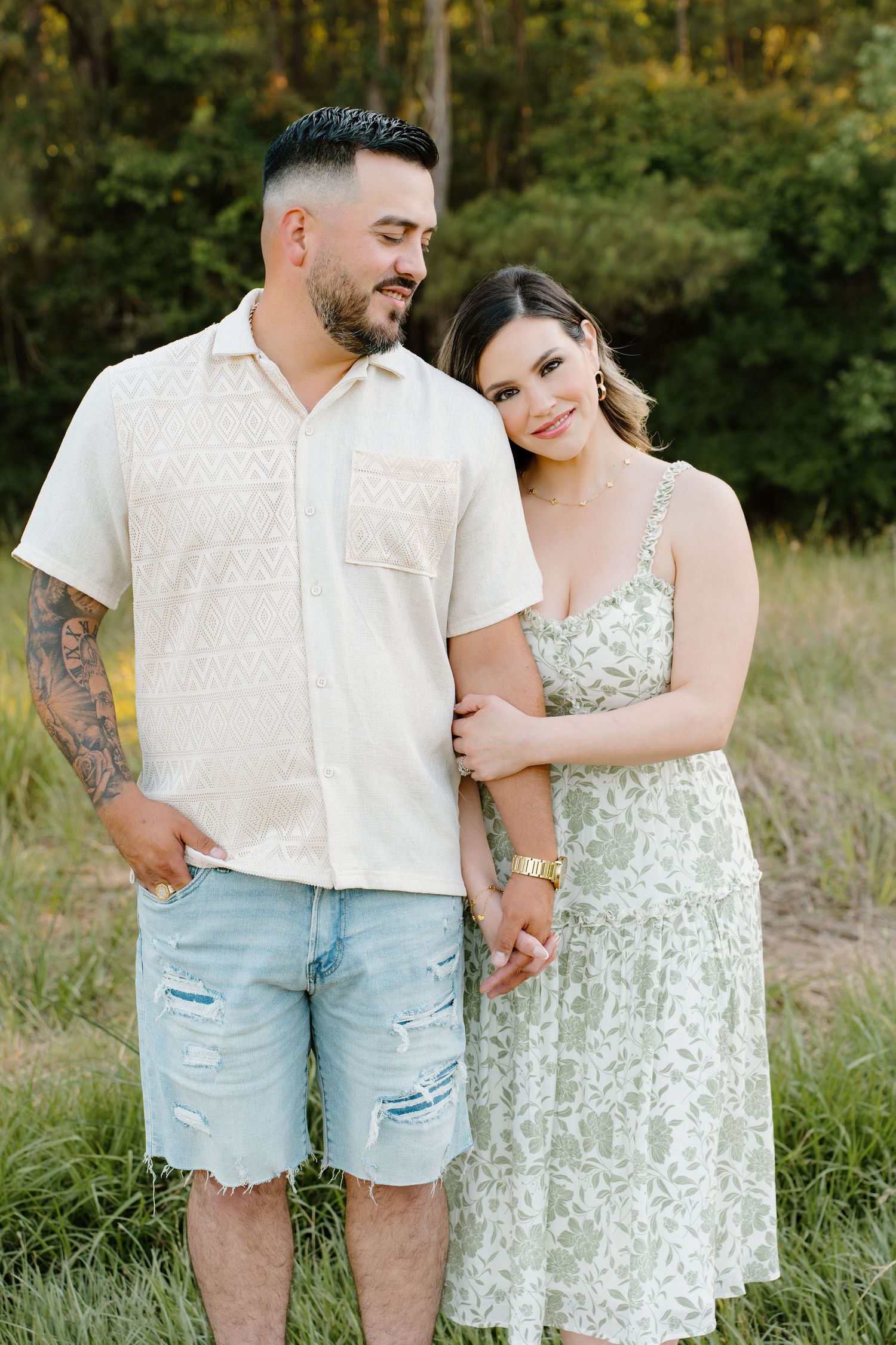 A couple poses outdoors in a grassy field, man in white shirt and denim shorts, woman in floral sundress.