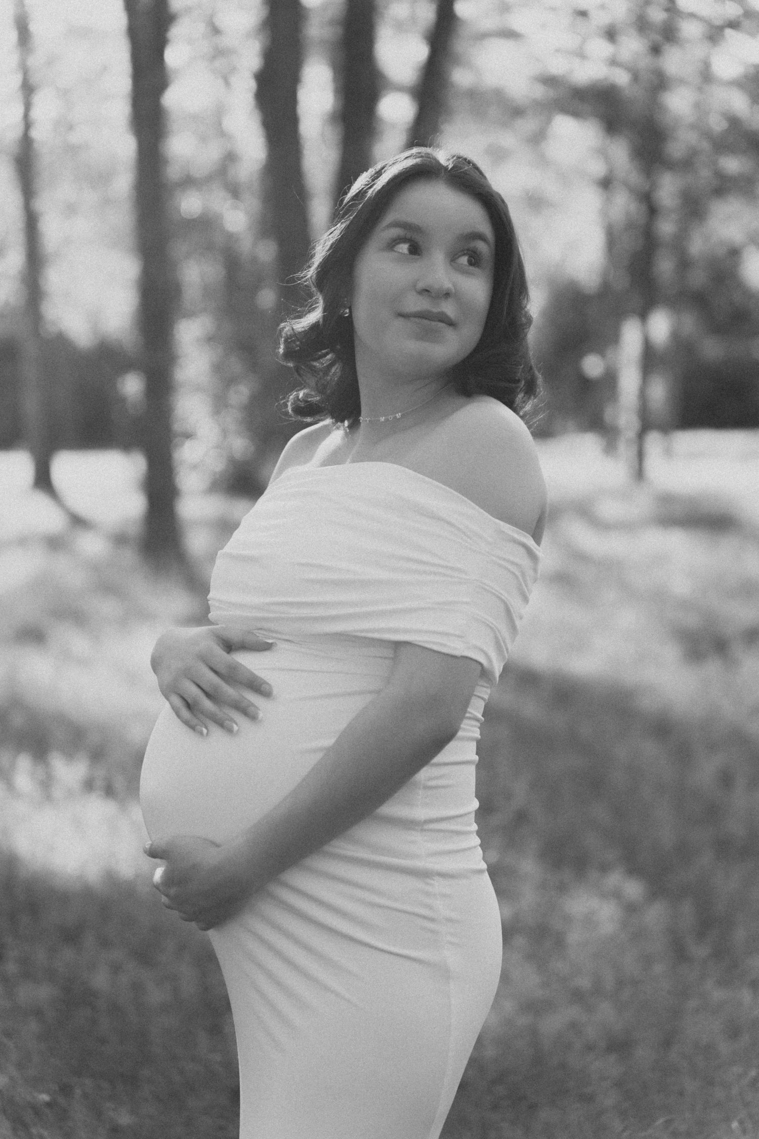 Black and white portrait of a pregnant woman in a white off-shoulder gown cradling her belly outdoors.