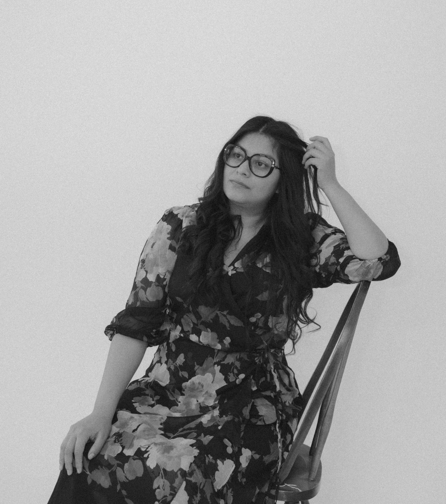 Black and white portrait of a woman in a floral dress sitting on a chair against a white wall.
