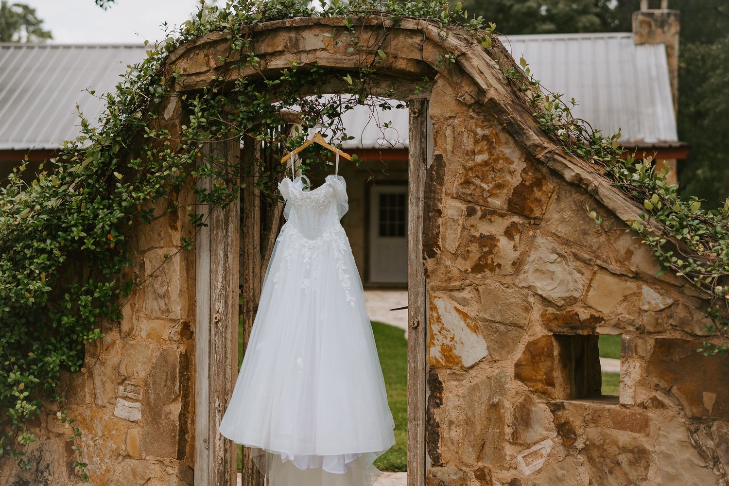 White wedding dress hanging on a rustic stone arch covered in greenery at an outdoor Texas Hill Country venue.