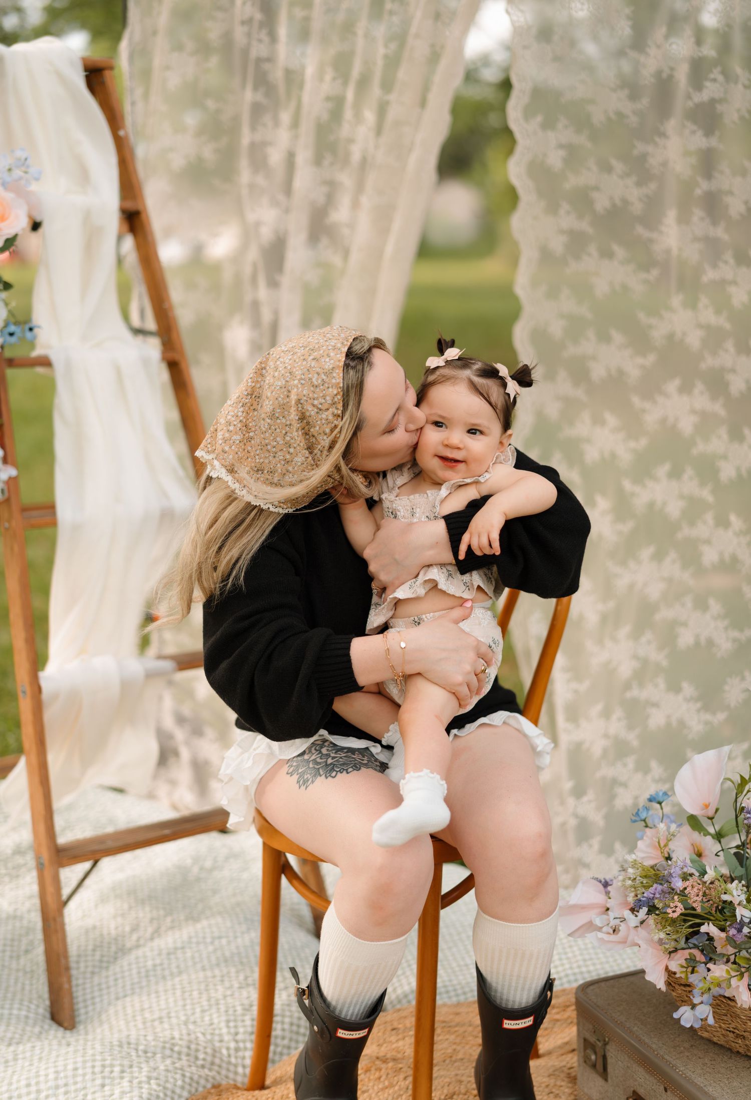 A mother kisses her smiling baby girl while seated on a wooden chair outdoors near sheer curtains.