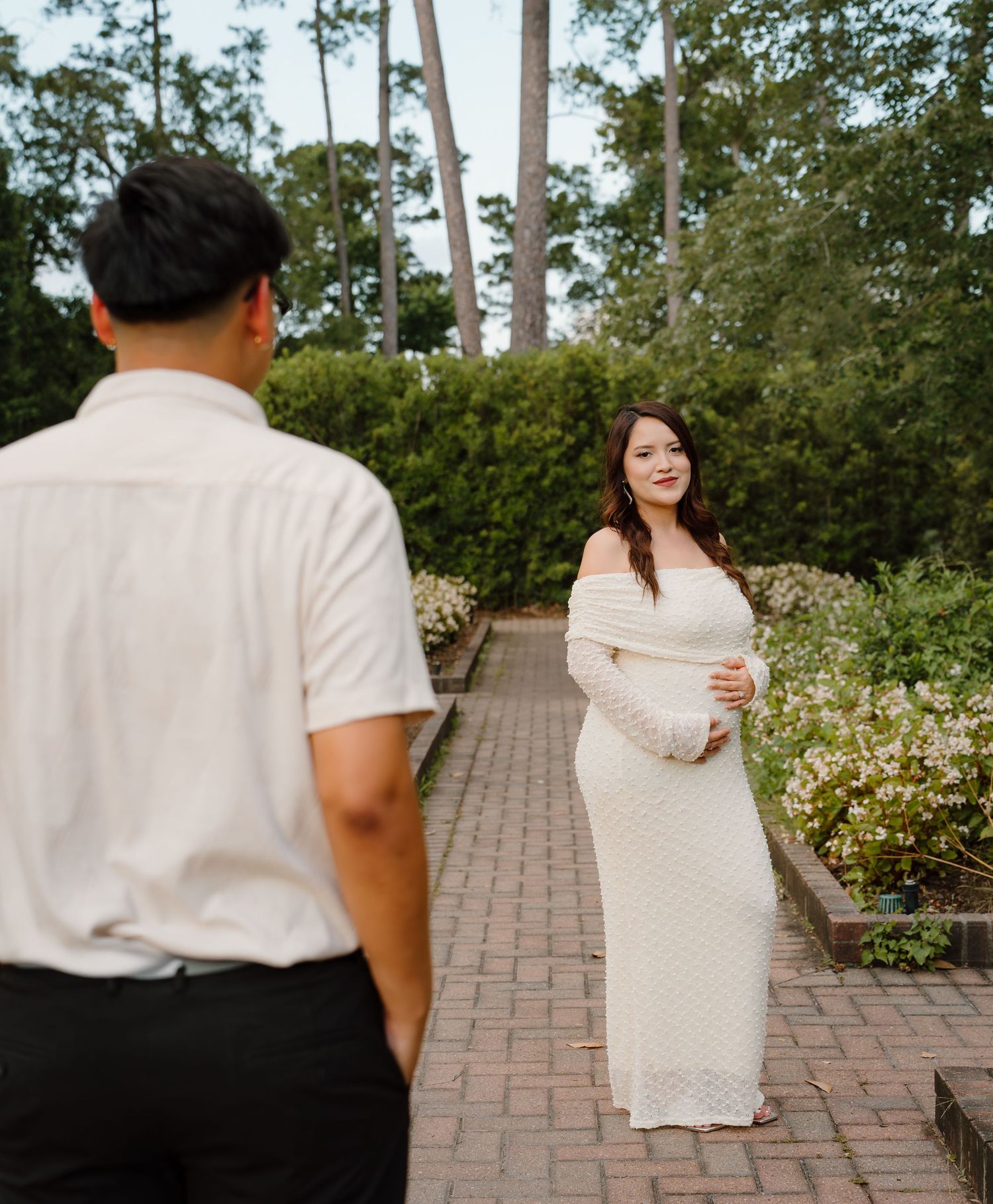 Pregnant woman in white off-shoulder knit dress poses in garden while partner looks on during maternity photoshoot.