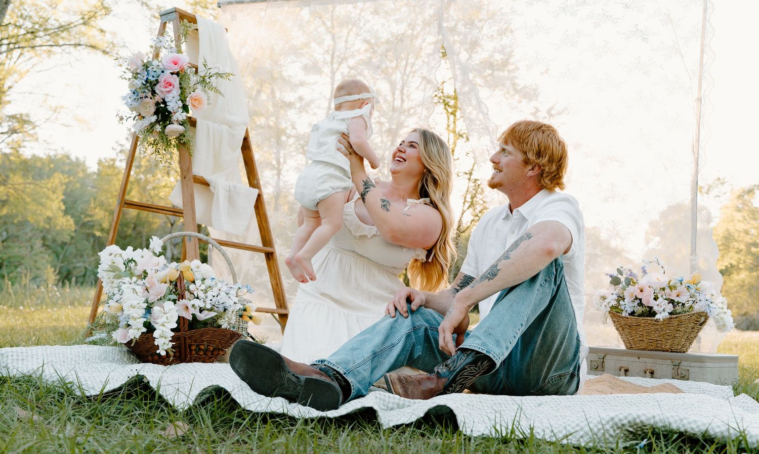 A happy family sits on a blanket outdoors with flowers, lifting their baby in warm sunlight.