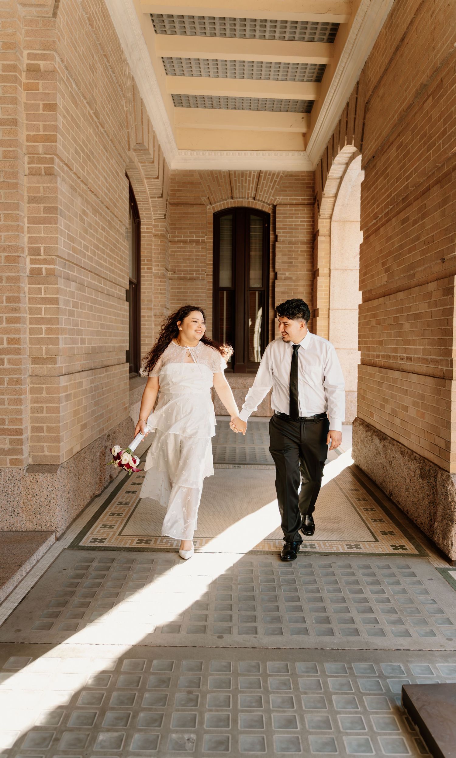 Newlywed couple walks hand in hand through sunlit brick archway corridor of historic building.