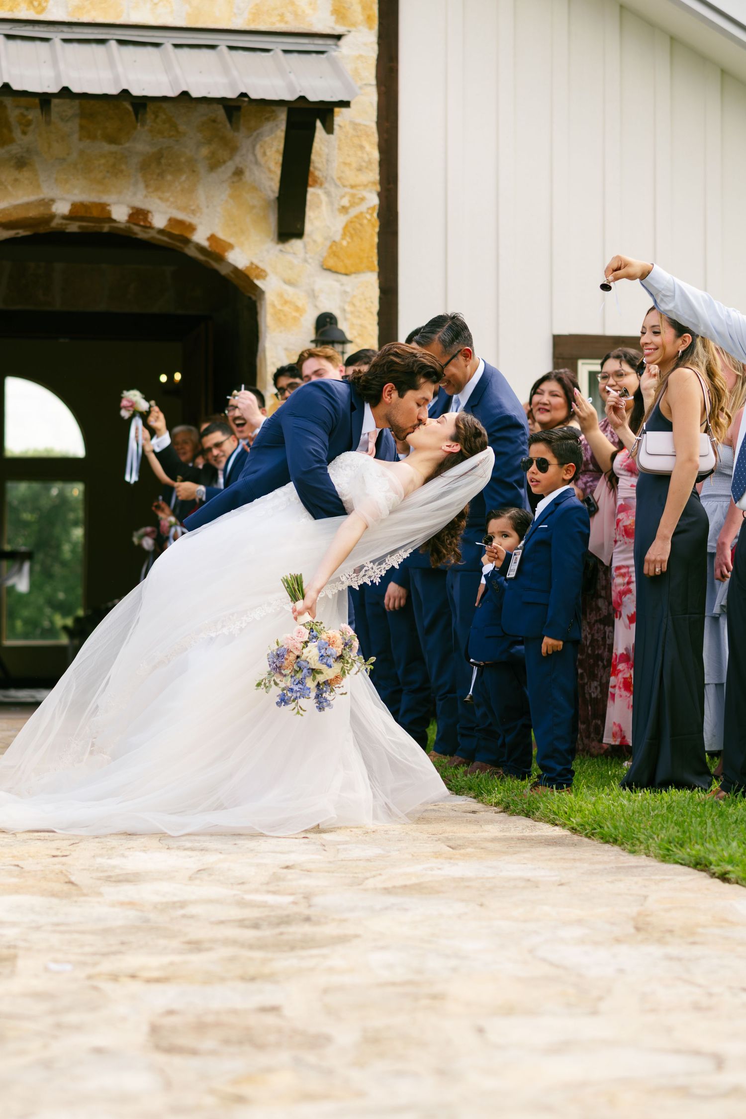 Groom dips bride for romantic kiss outside stone chapel surrounded by cheering wedding party guests.