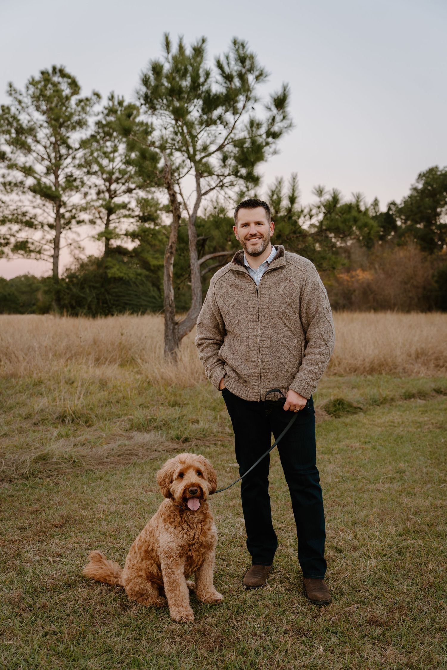 A smiling man in a tan sweater stands with a golden doodle dog in a grassy field at dusk.