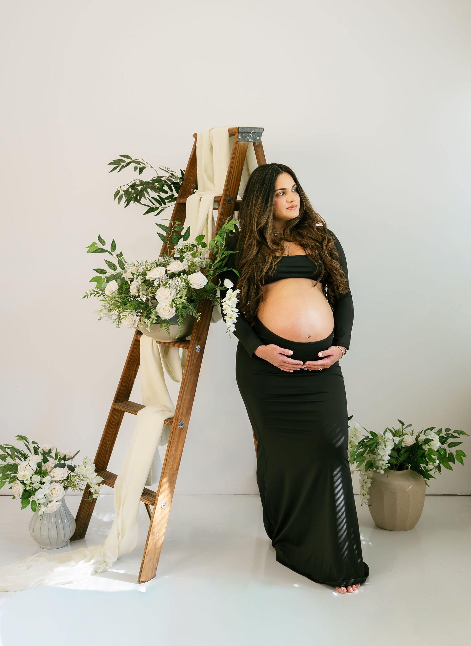 Pregnant woman in dark crop top and skirt poses by rustic ladder with white florals.