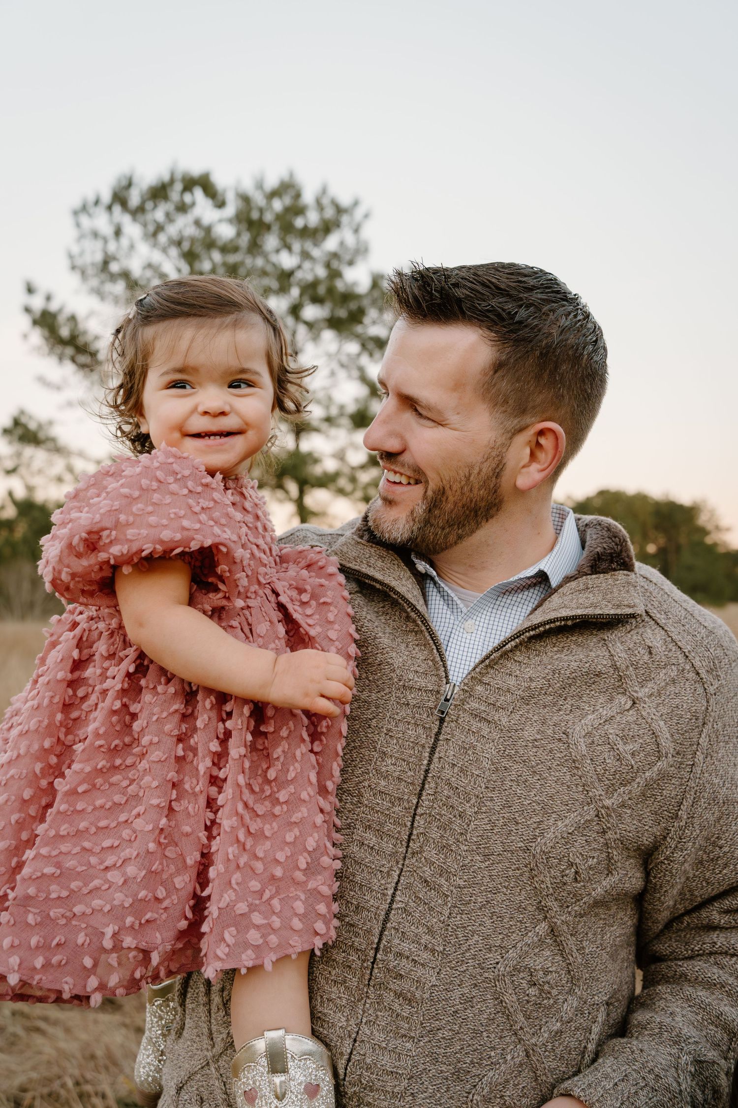 A smiling man in a tan sweater holds a happy toddler girl in a pink dress outdoors at sunset.