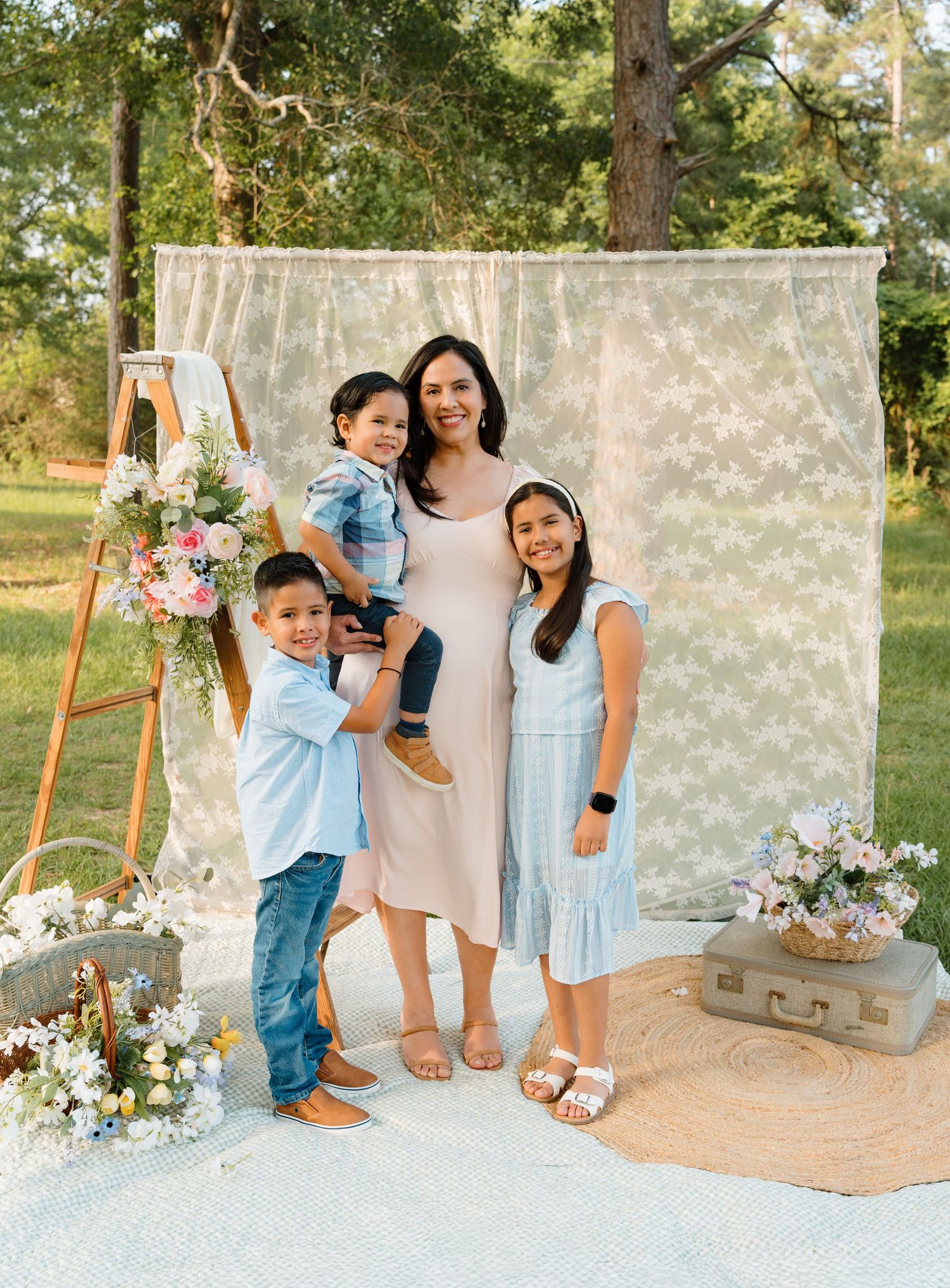 A mother poses outdoors with her three children in front of a lace backdrop with floral decorations.