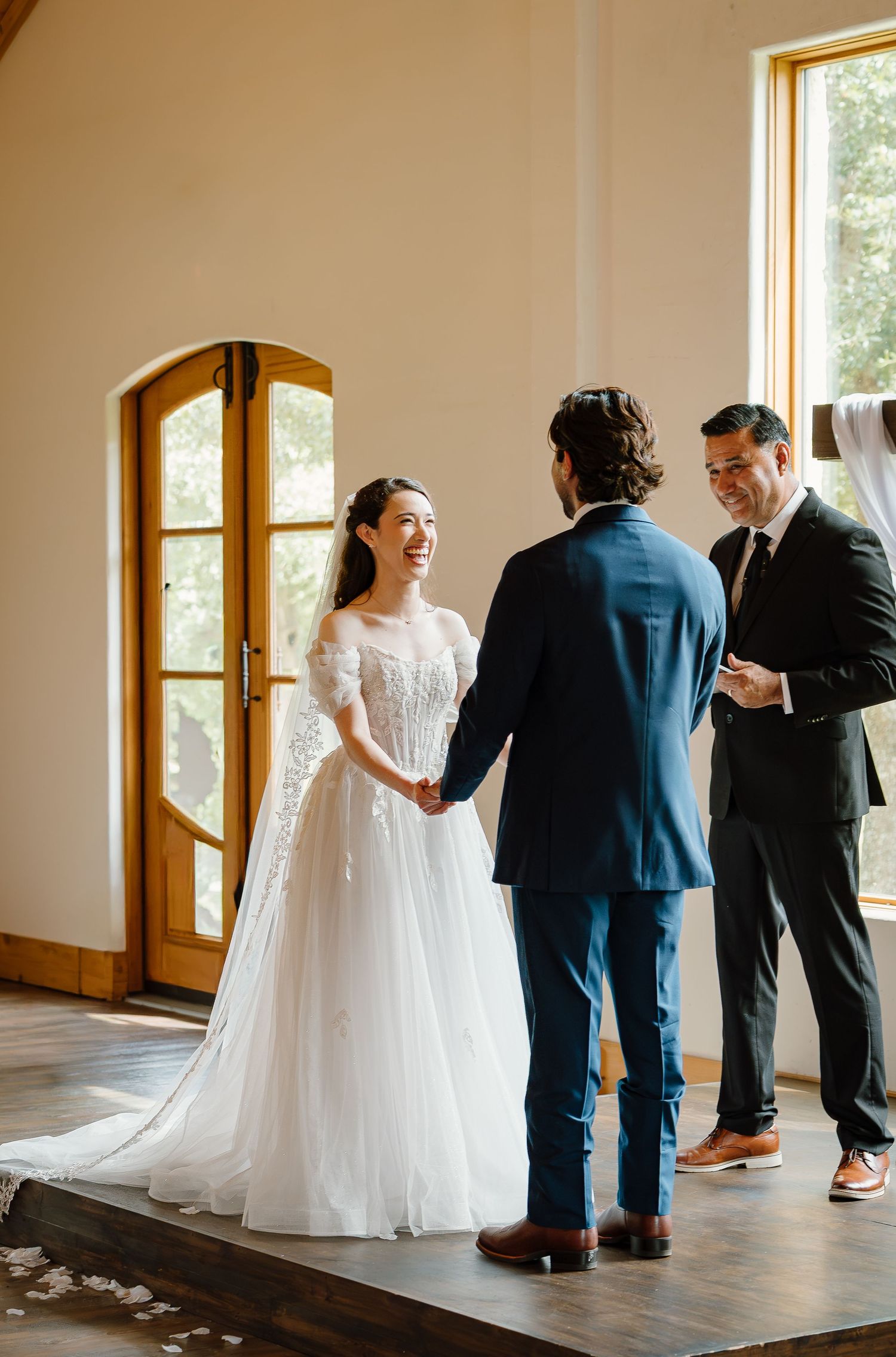 Bride in white gown laughing with groom in navy suit during indoor wedding ceremony near wooden doors.