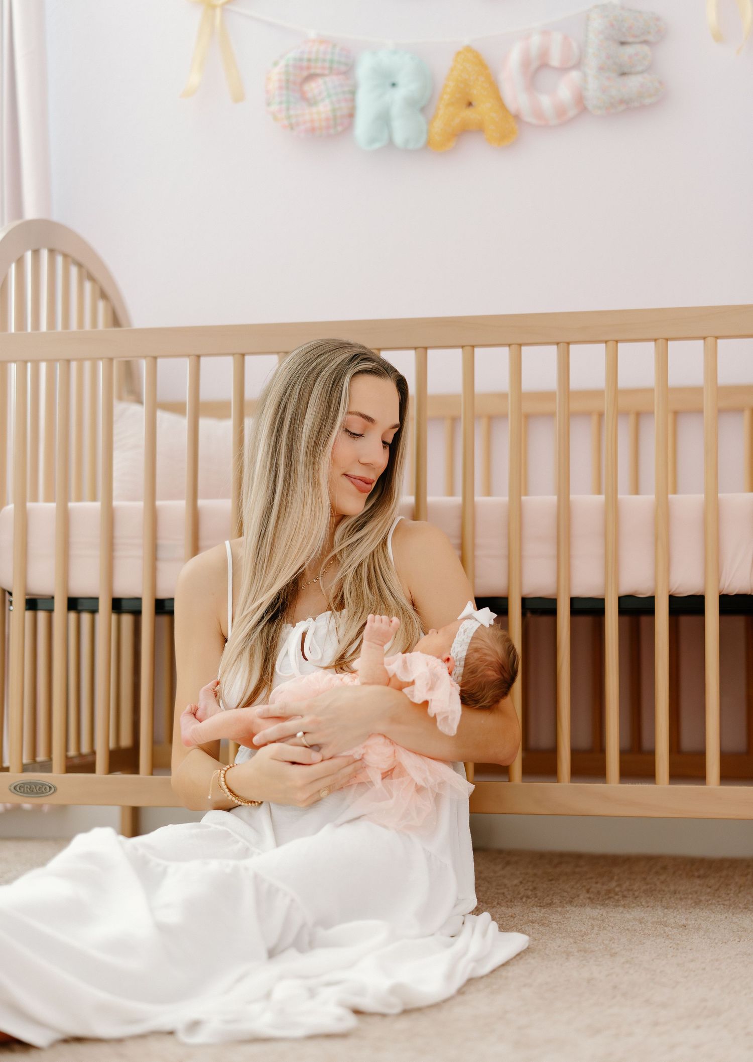 A mother sits on the floor cradling her newborn baby in a nursery with a wooden crib and colorful 'GRACE' name banner.