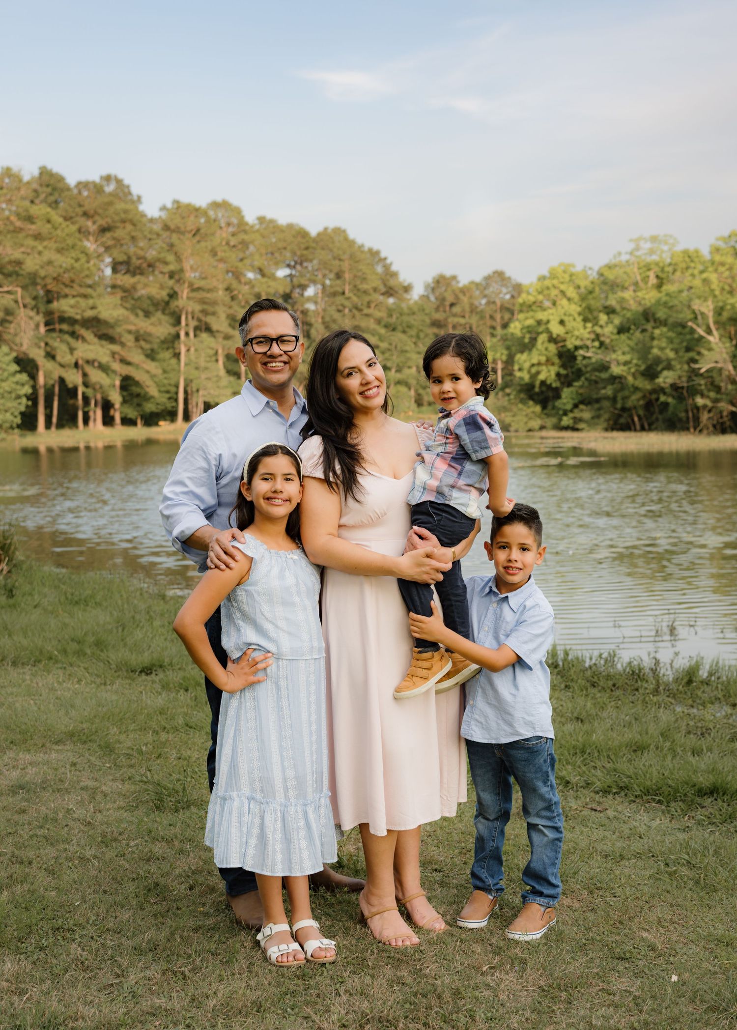 Family of five smiling outdoors by a serene lake, surrounded by lush green trees on a sunny day.