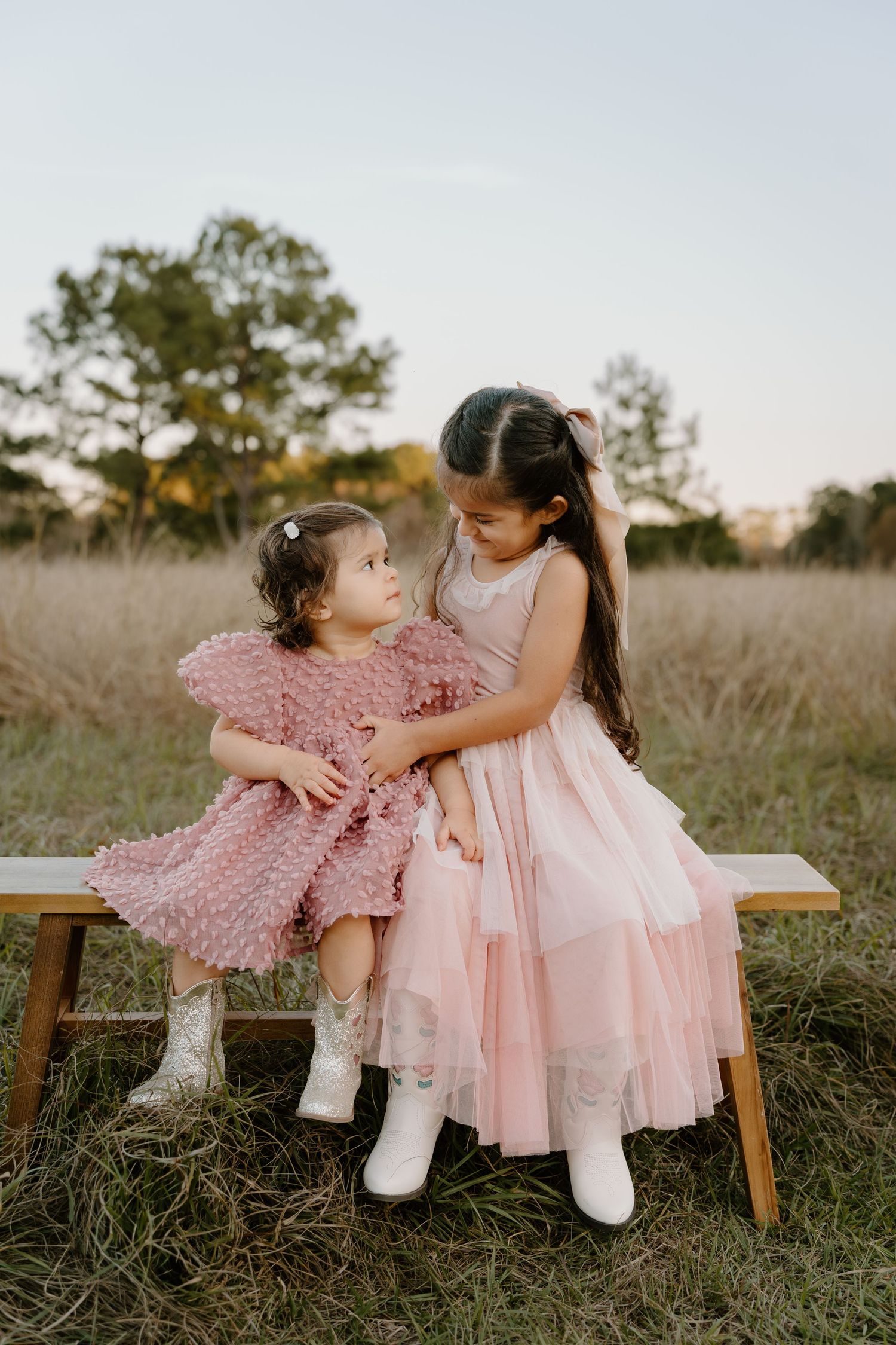 Two young girls in matching pink dresses sit on a wooden bench in a field, sharing a sweet sisterly moment.