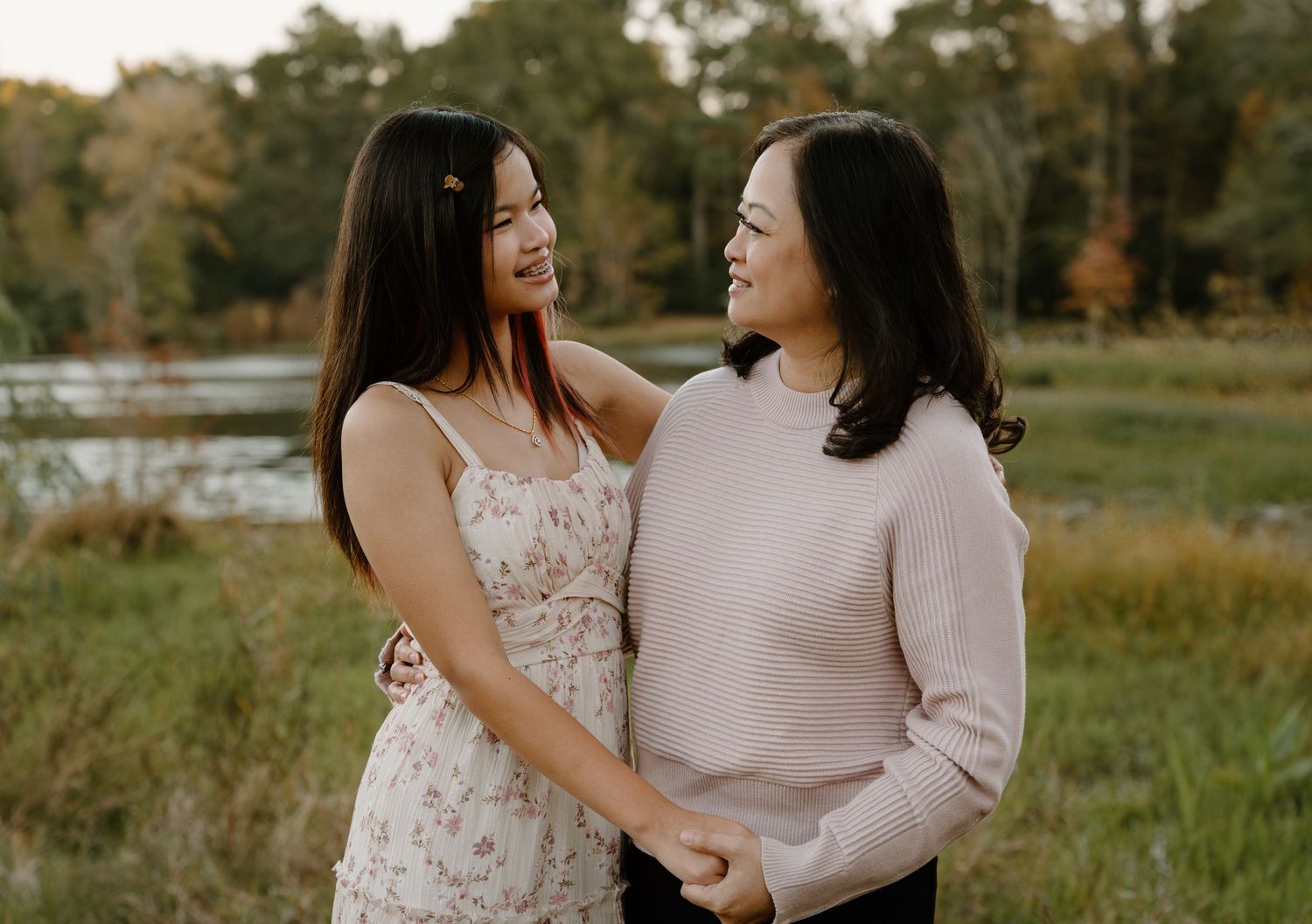 Two women smiling and holding hands outdoors in a scenic park setting with autumn foliage in the background.