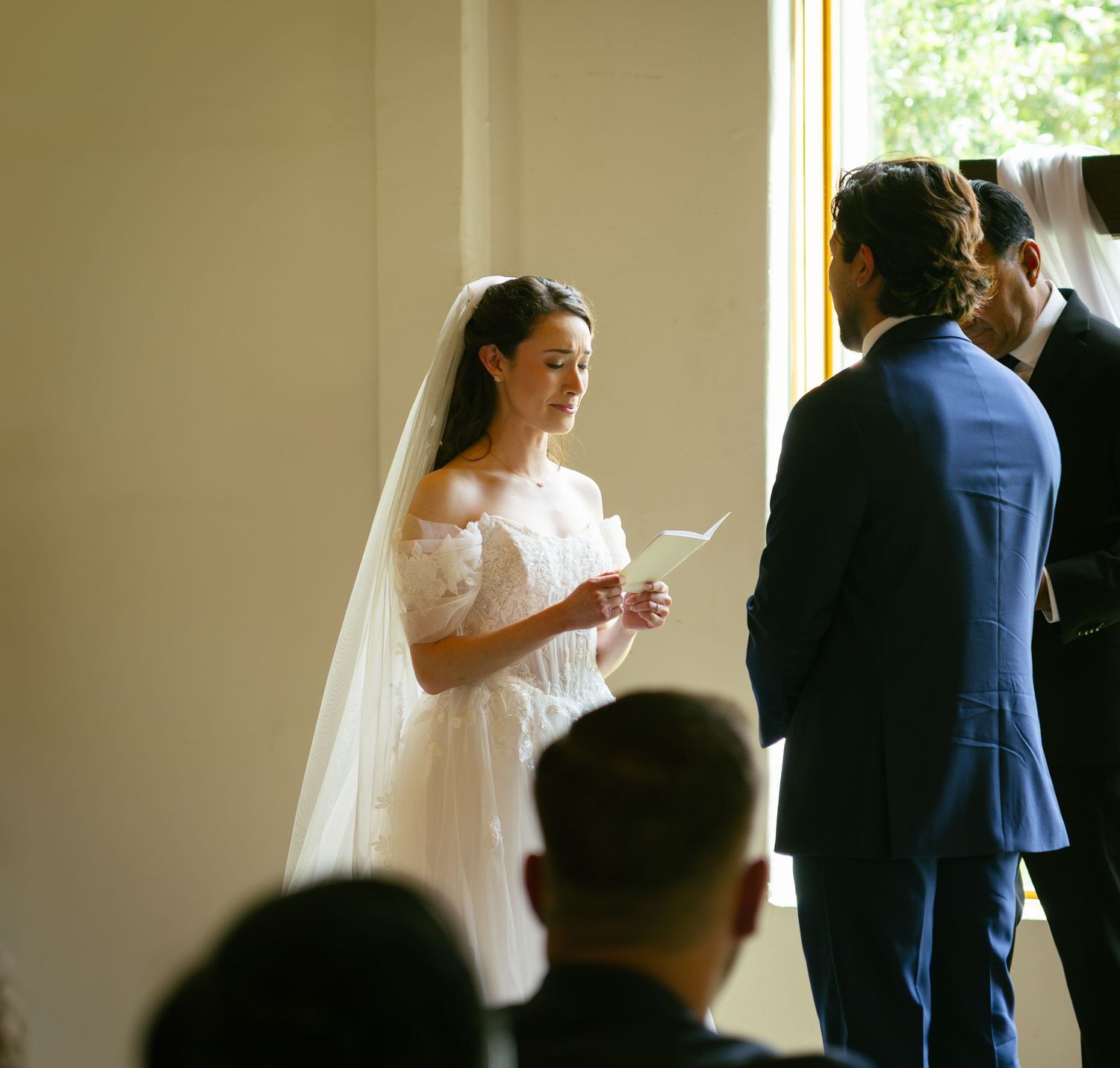 Bride in white lace off-shoulder gown reads vows to groom in navy suit during indoor wedding ceremony.
