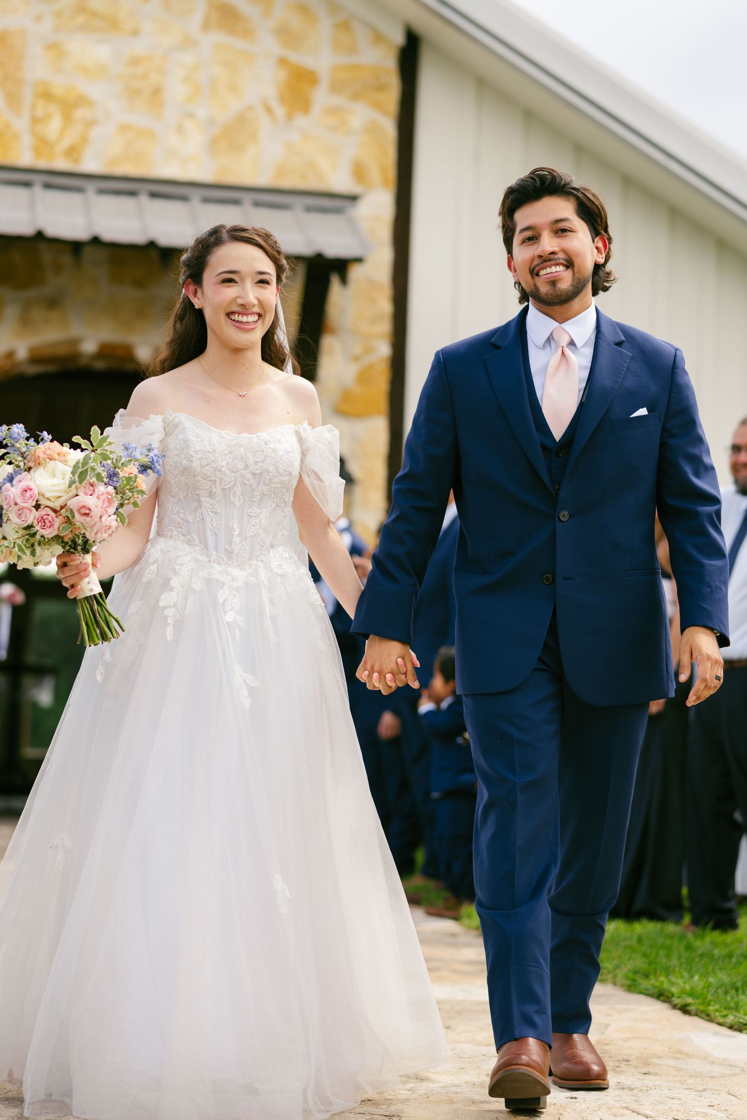 Smiling newlyweds walking hand in hand after ceremony, bride in white gown holding floral bouquet.