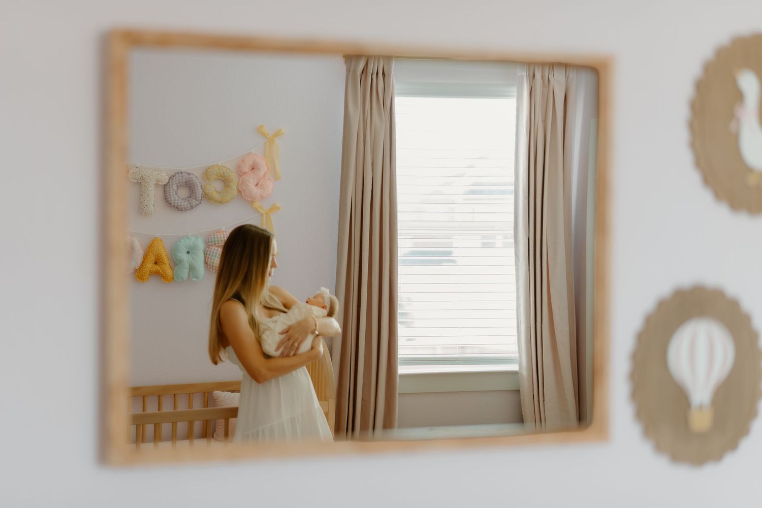 Mother holding newborn baby in nursery room, reflected in wooden framed mirror with colorful letter banner.