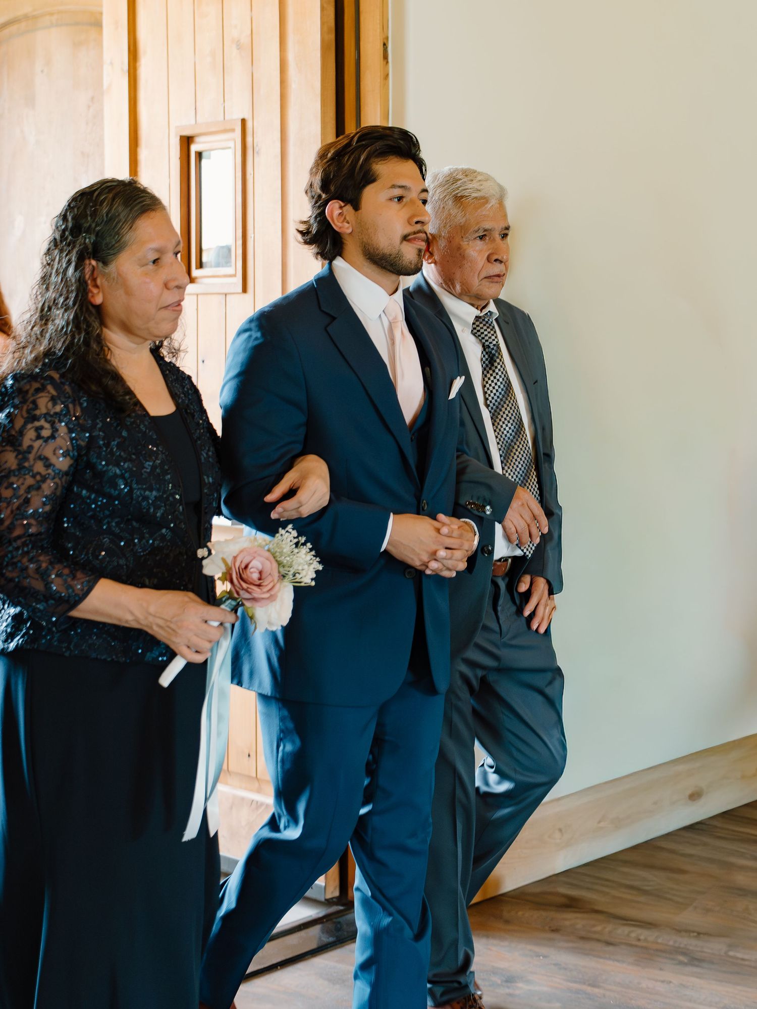 A groom in a navy suit is escorted down the aisle by his parents during a wedding ceremony.