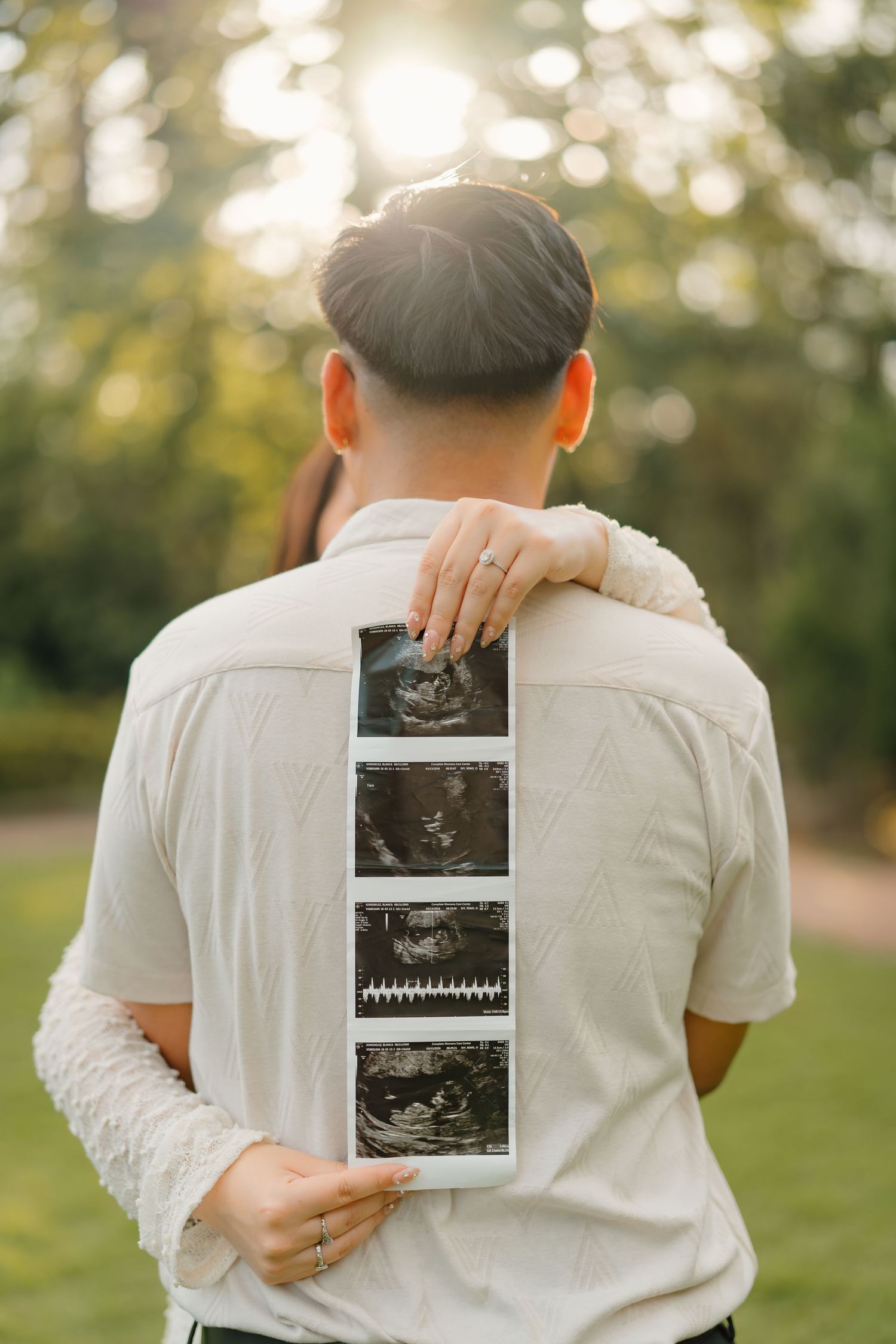 Couple embracing outdoors holding ultrasound photos, pregnancy announcement in sunlit park setting.