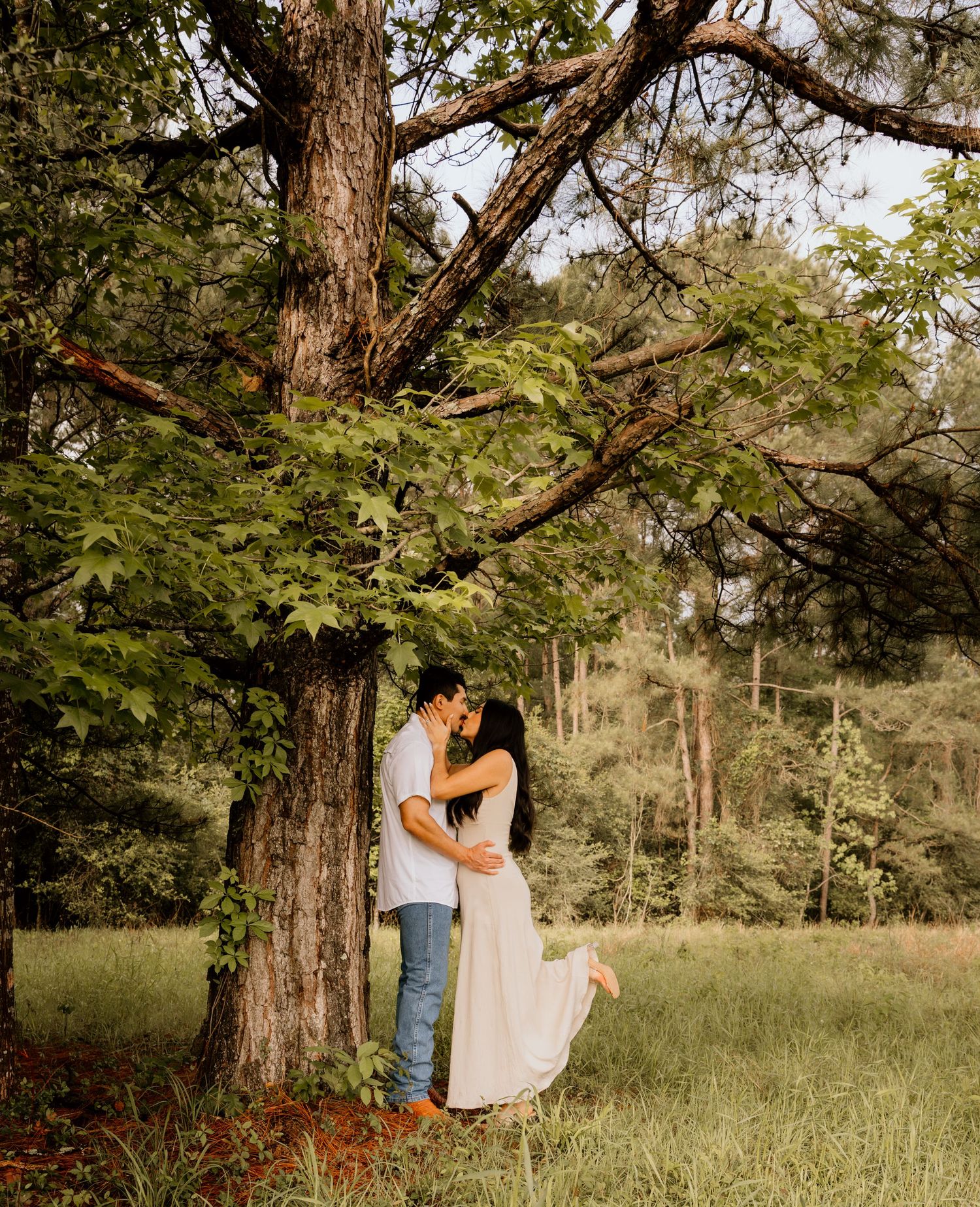 Couple sharing a kiss under a large oak tree in a lush green meadow, woman in flowing white dress.