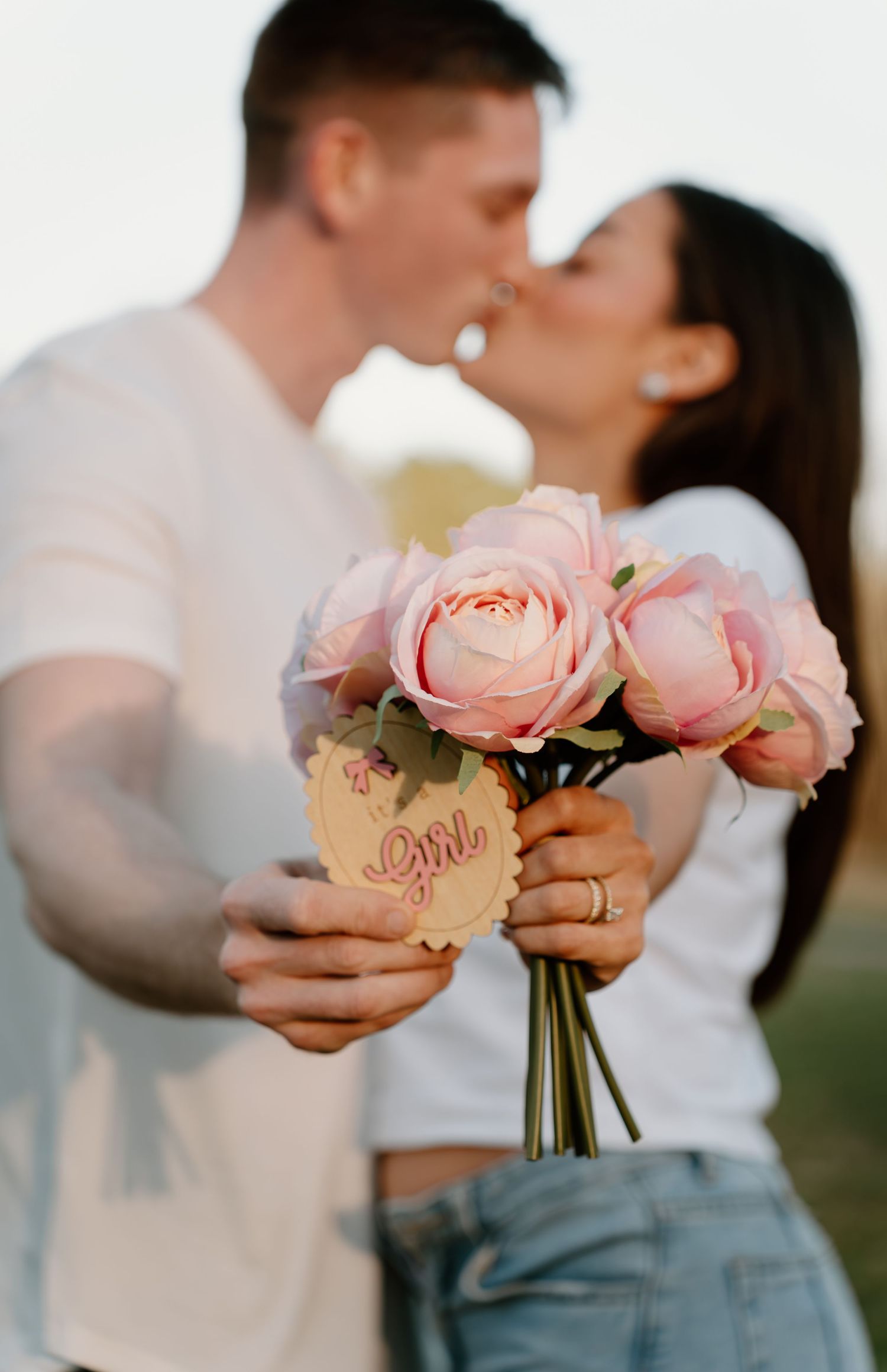 Couple kissing while holding pink peony bouquet with a girl gender reveal tag.