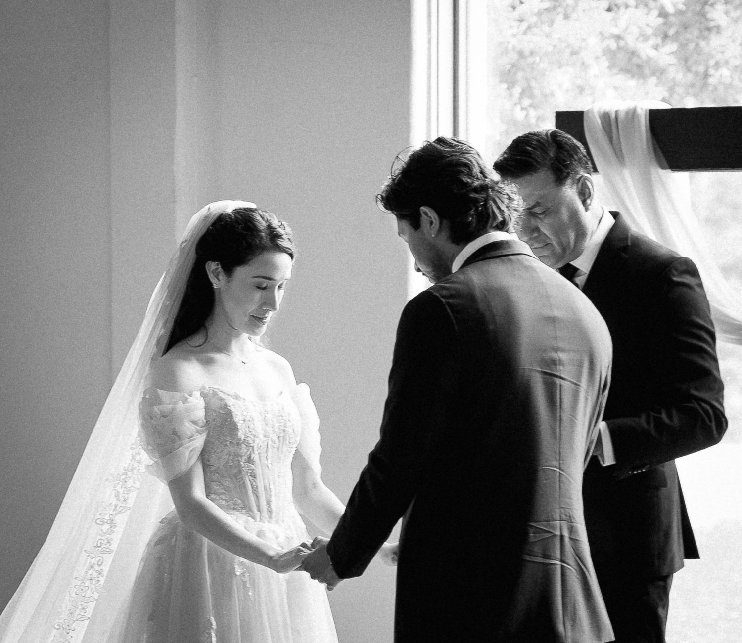 Black and white wedding ceremony photo of bride in lace gown holding hands with groom before officiant.
