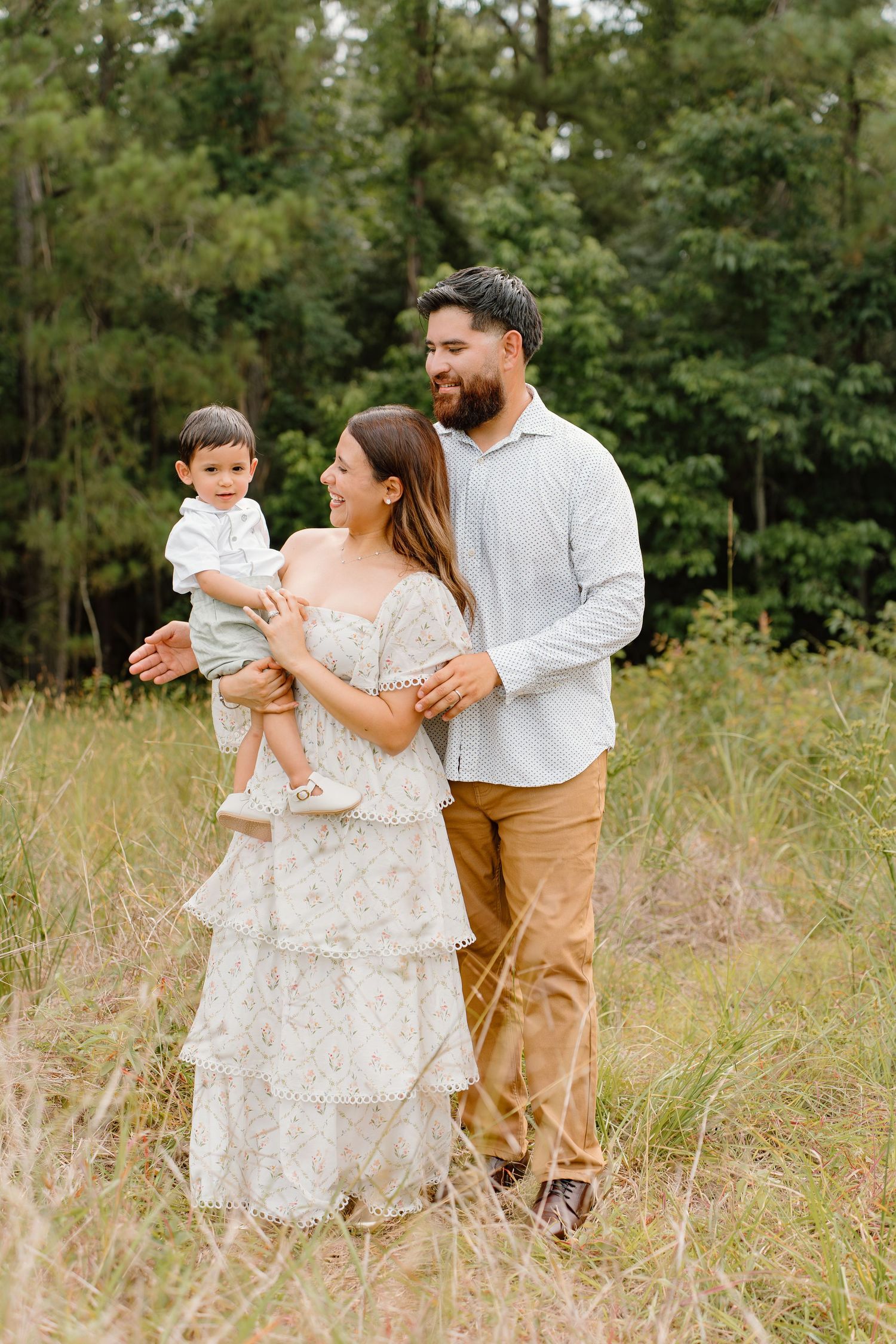 A happy young family poses outdoors in a lush green field, with parents holding their toddler in matching neutral outfits.