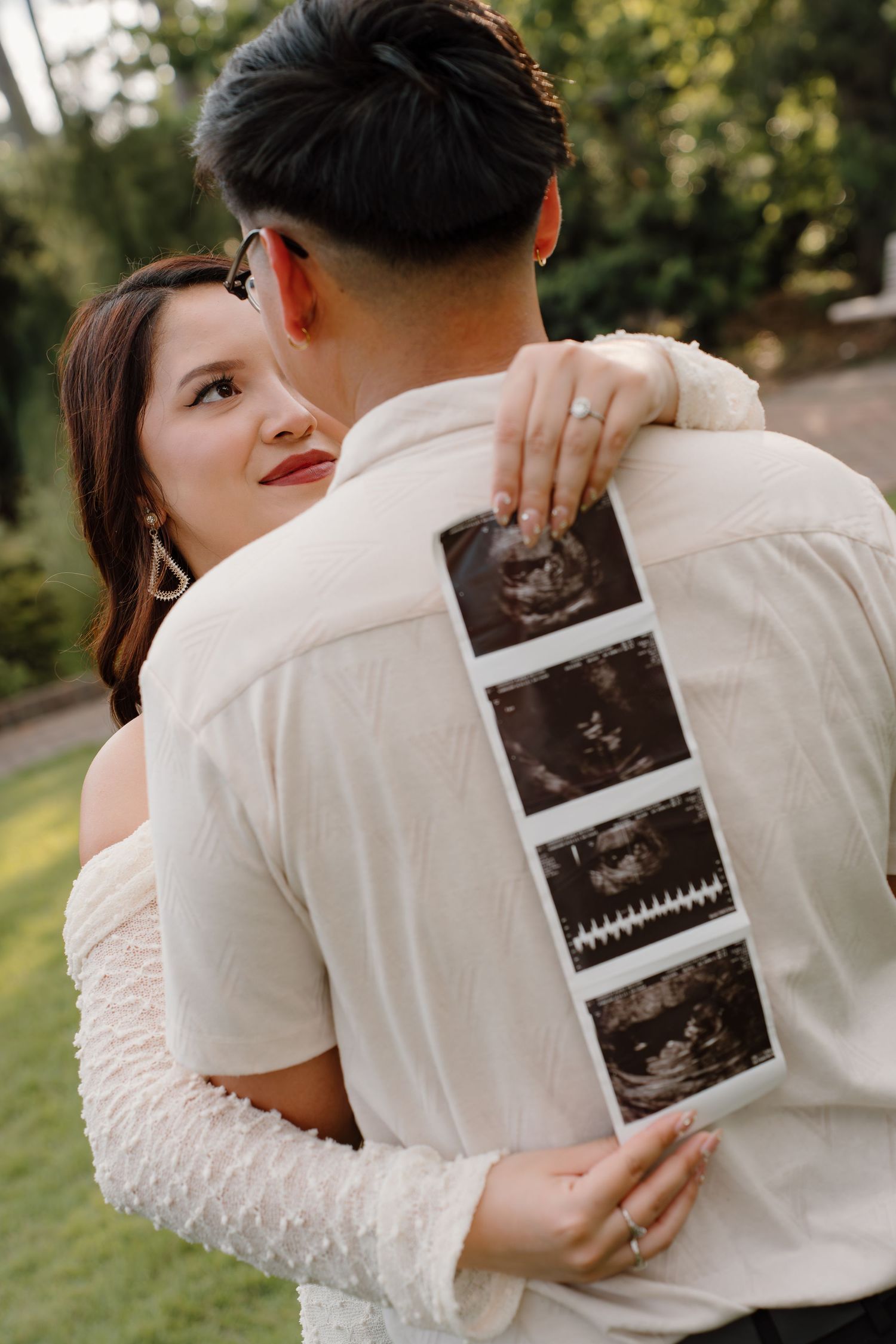 Couple embracing outdoors, woman holding ultrasound photos behind man's back, pregnancy announcement photo.