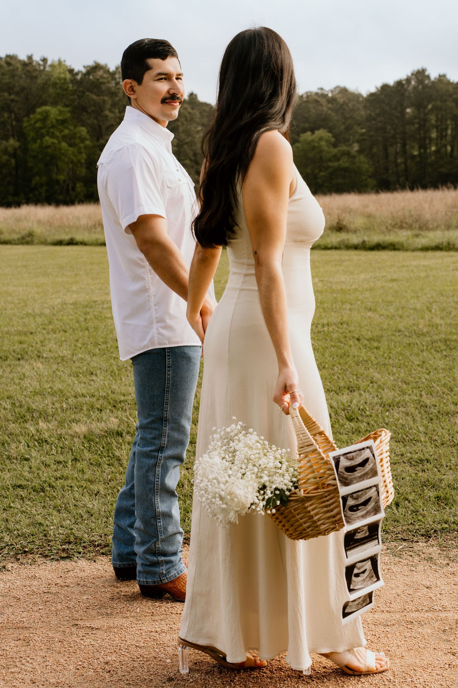 Couple holding hands in field, woman in white dress carrying basket with flowers and ultrasound photos, pregnancy reveal.