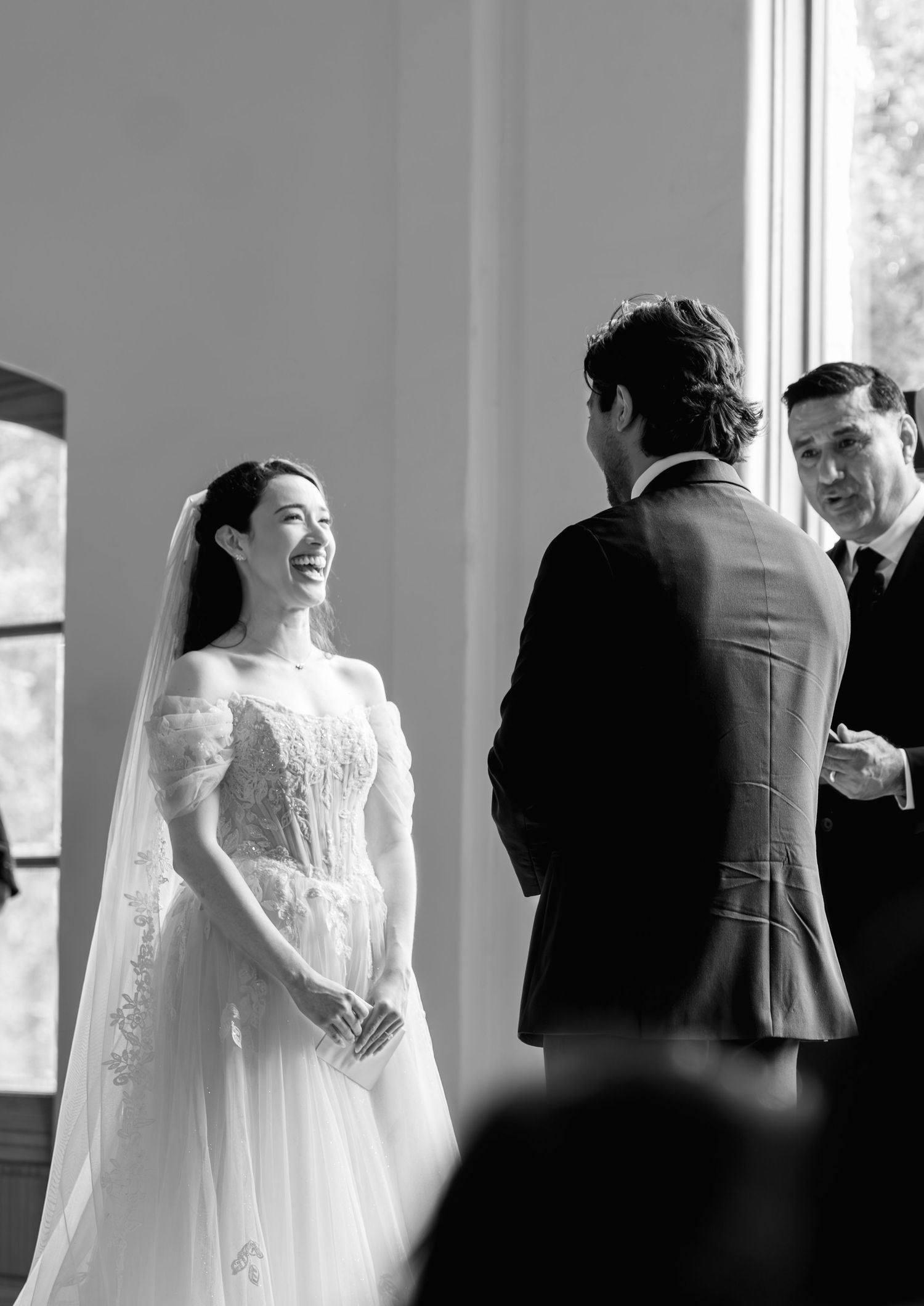 Black and white photo of a smiling bride in a lace gown exchanging vows with groom at wedding ceremony.