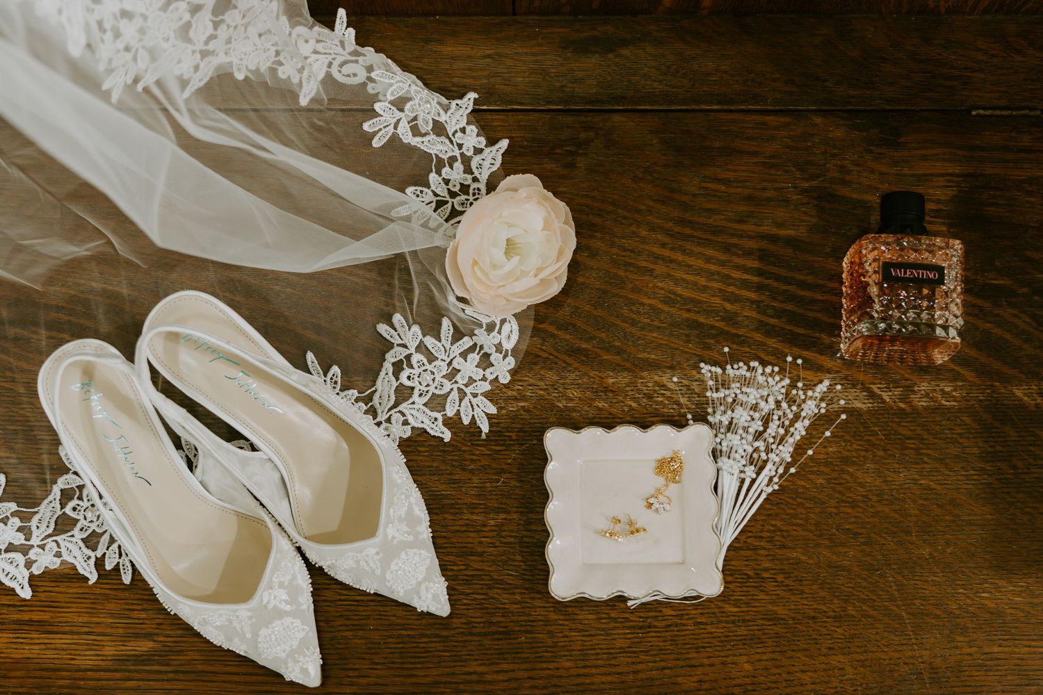 Elegant bridal flat lay featuring lace heels, veil, floral hair piece, perfume bottle, and delicate jewelry on rustic wood.