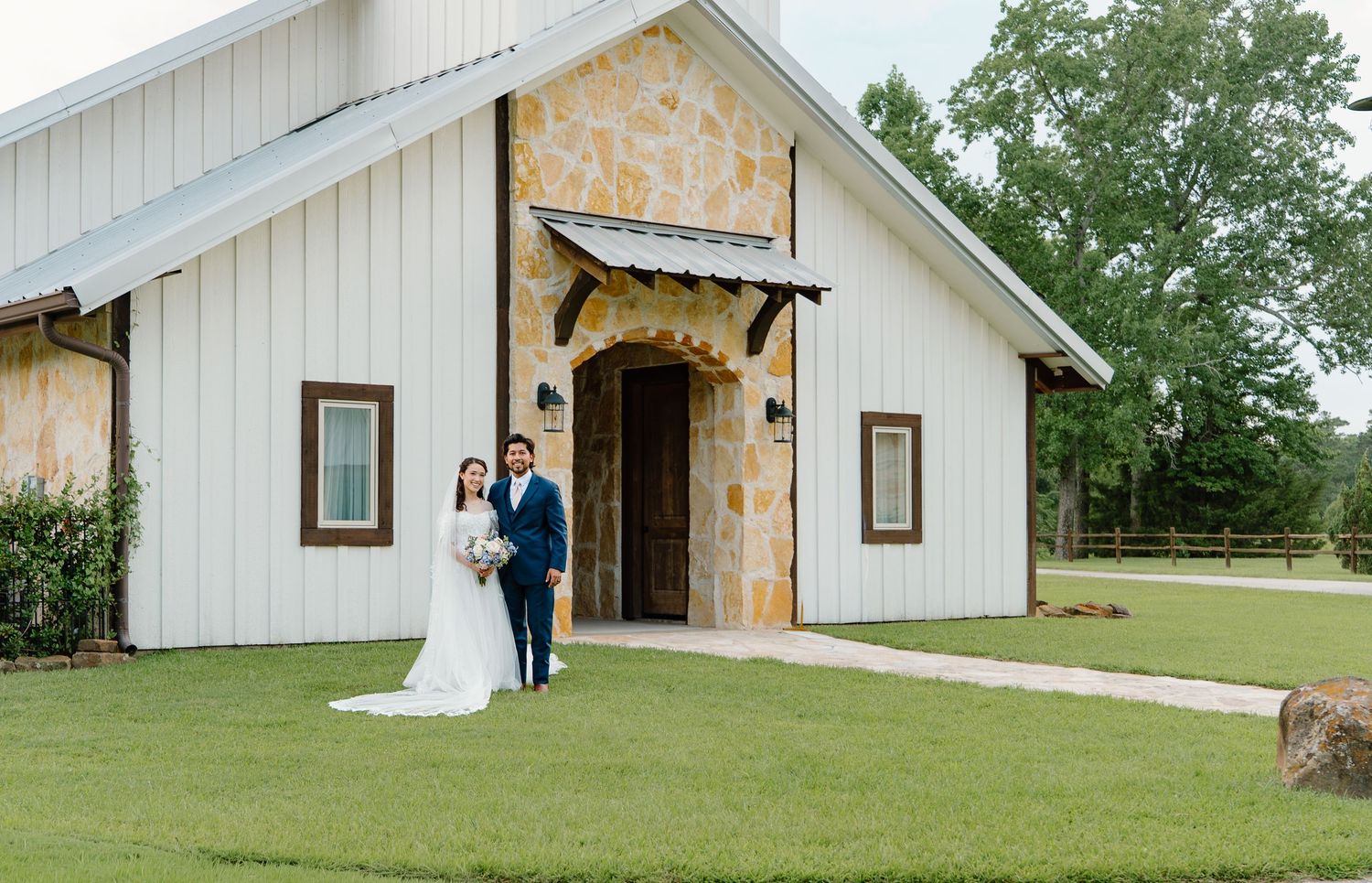 Bride and groom pose outside a white barn wedding venue with stone archway entrance surrounded by lush green trees.
