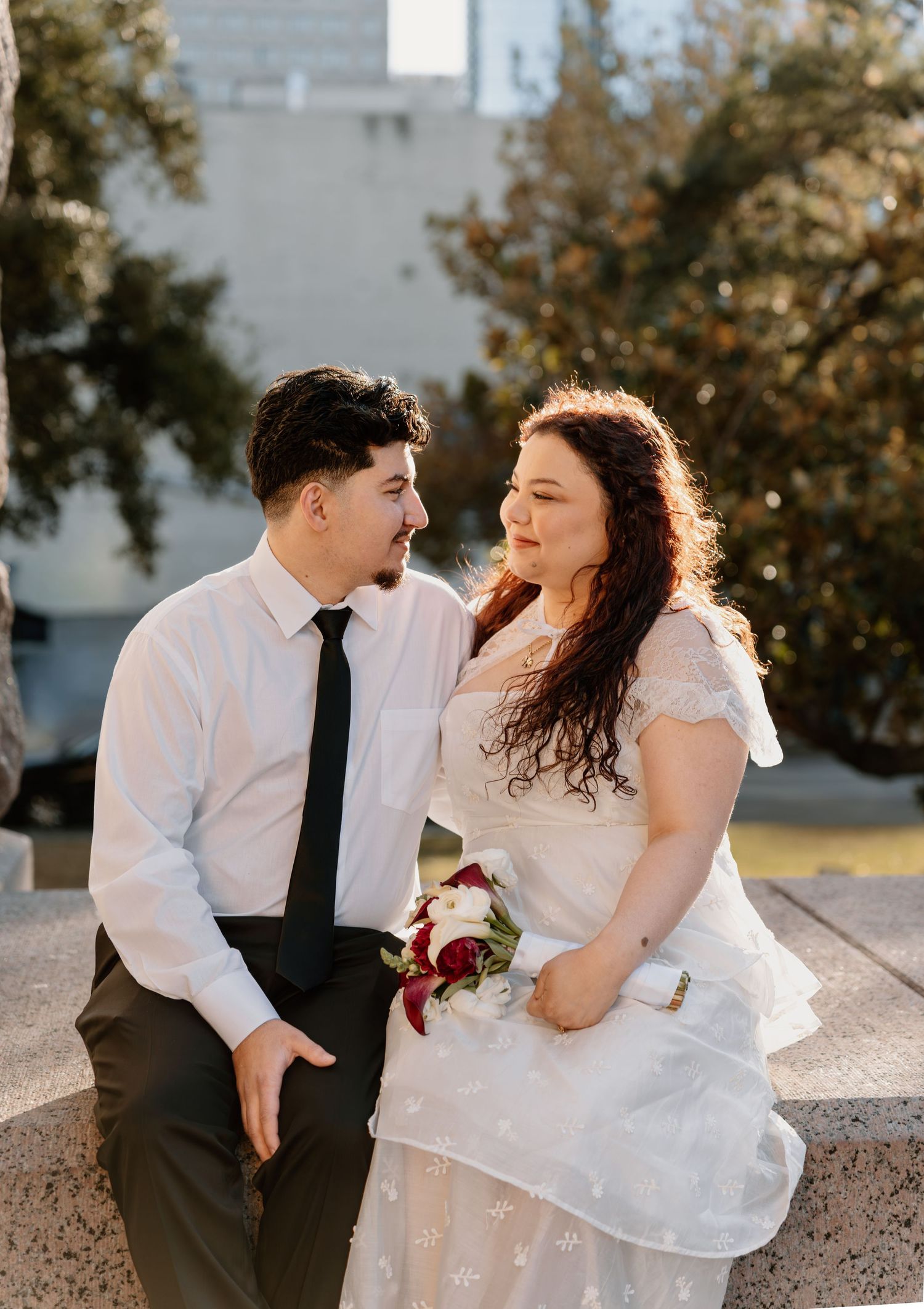 Bride and groom sit on stone ledge smiling at each other, bride holds red and white bouquet outdoors.