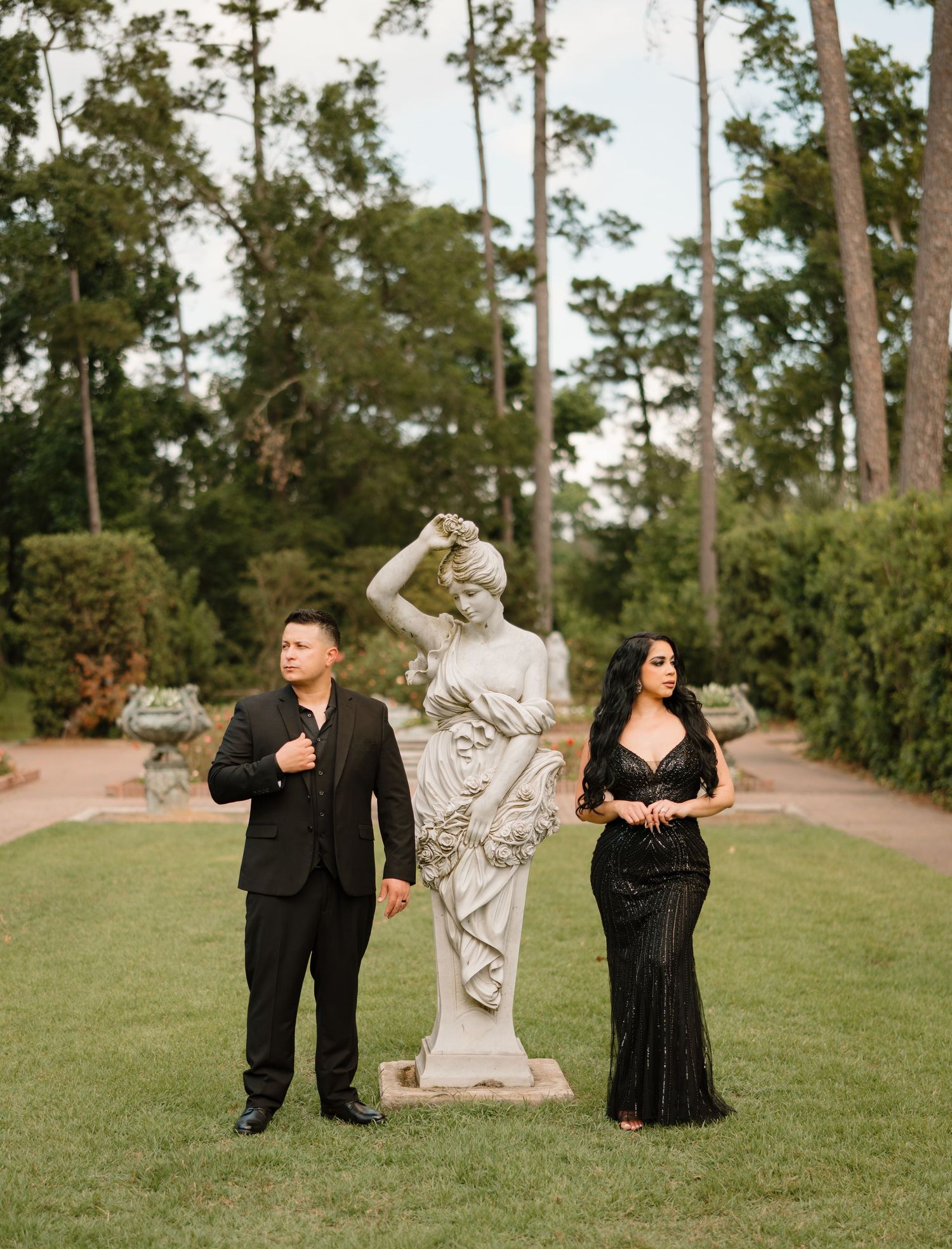 Couple in elegant black formal attire posing beside a classical white statue in a lush garden setting.