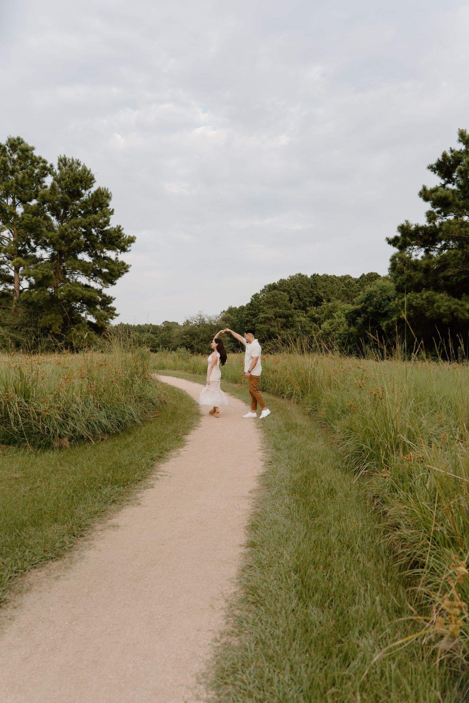 Couple dancing on a winding path surrounded by lush green fields and trees under a cloudy sky.