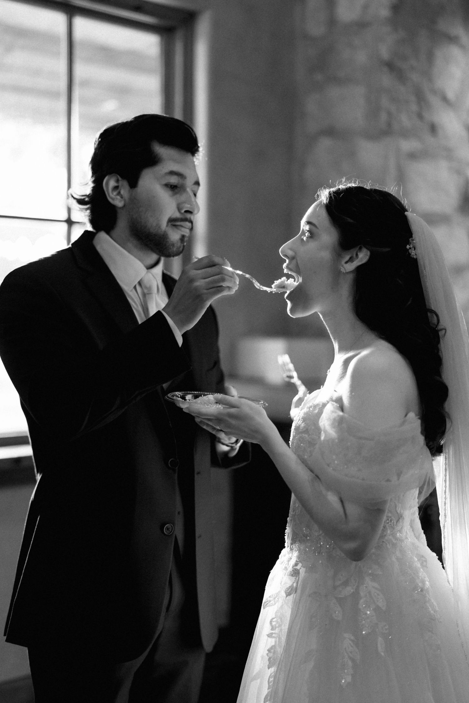 Black and white photo of bride and groom feeding each other wedding cake in a rustic venue.