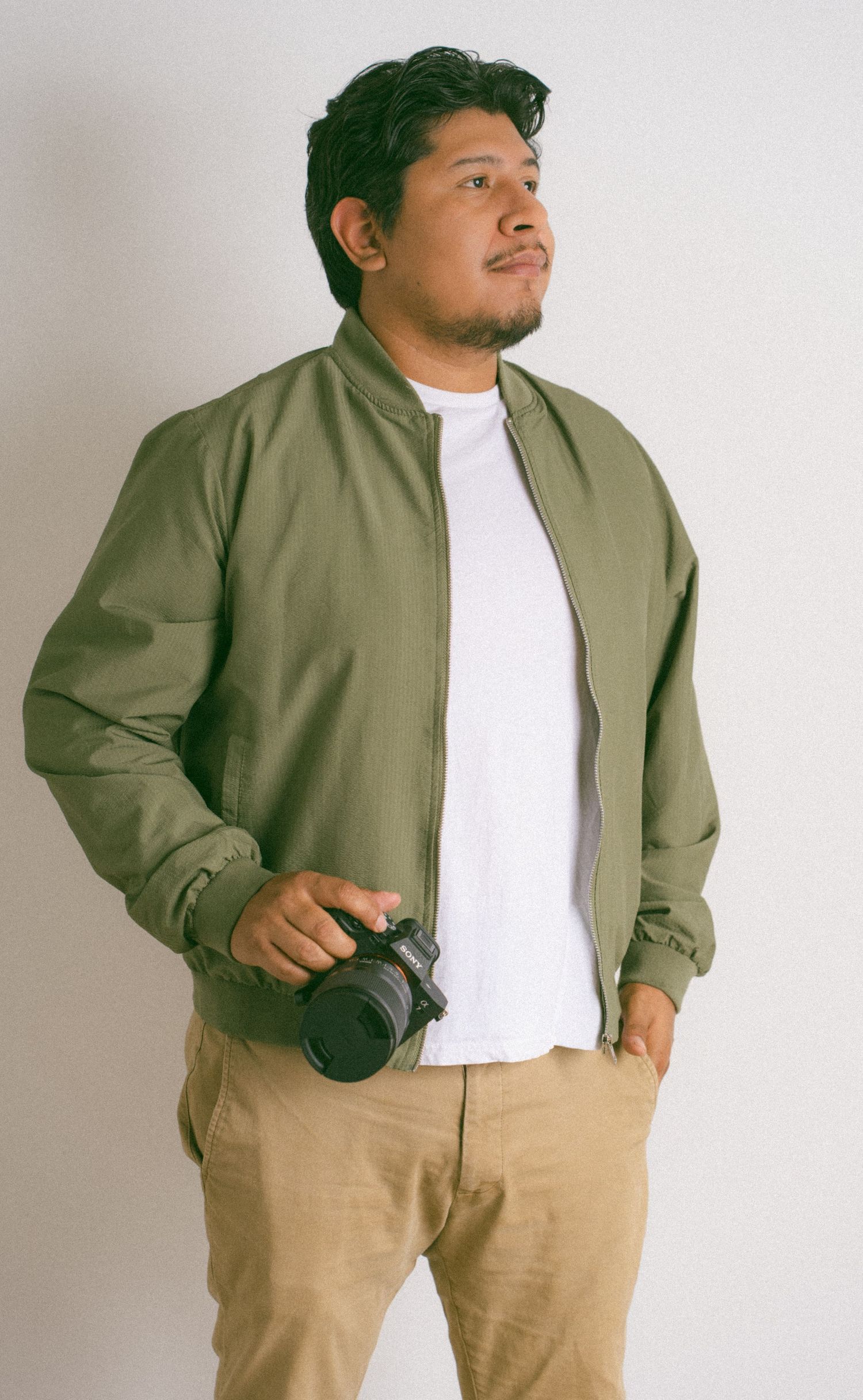 A male photographer in an olive bomber jacket holds a camera while posing against a white wall.